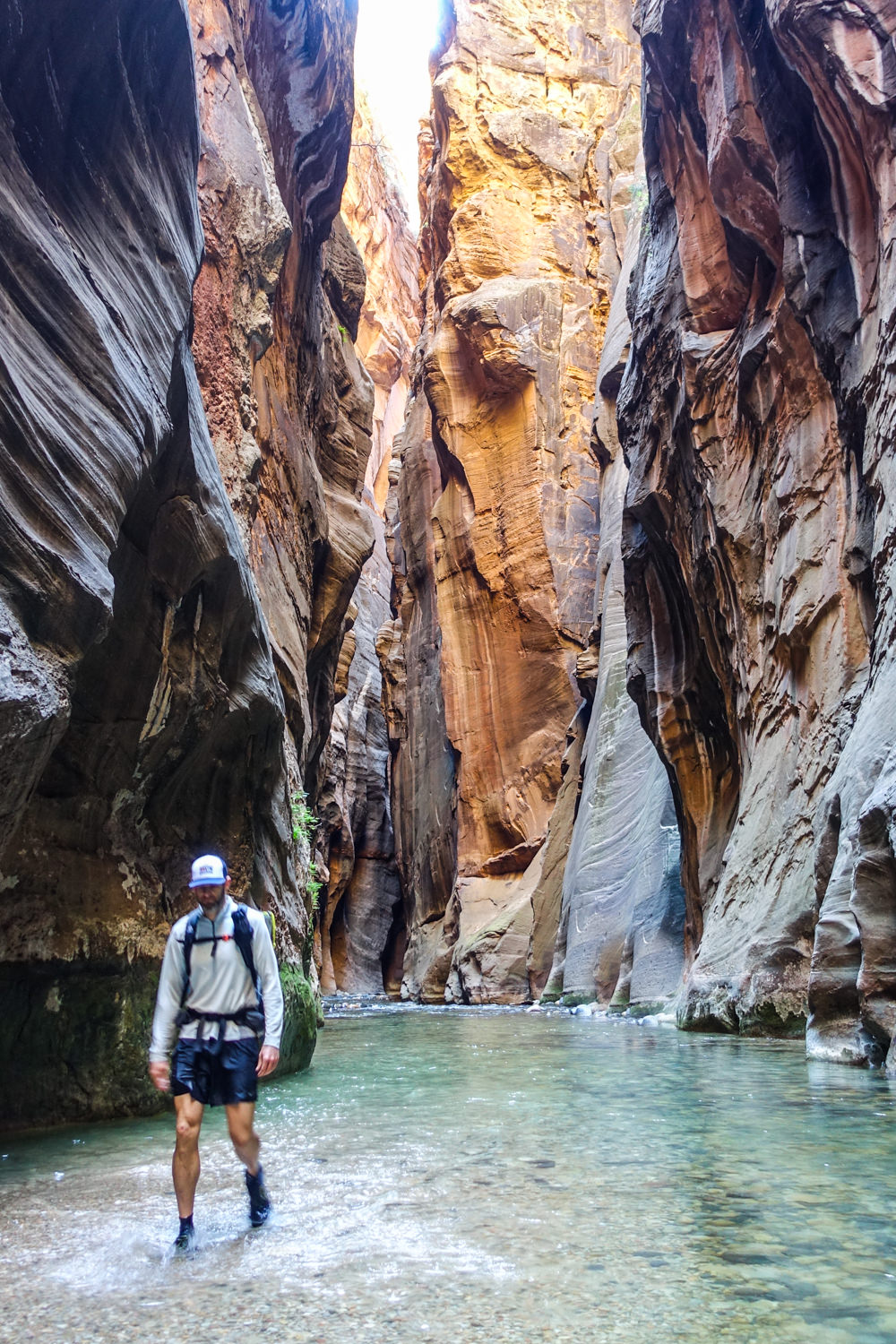 the narrow undulating orange and tan stiped walls of the narrows slot canyon with a hiker walking through the shallow river that carves the walls