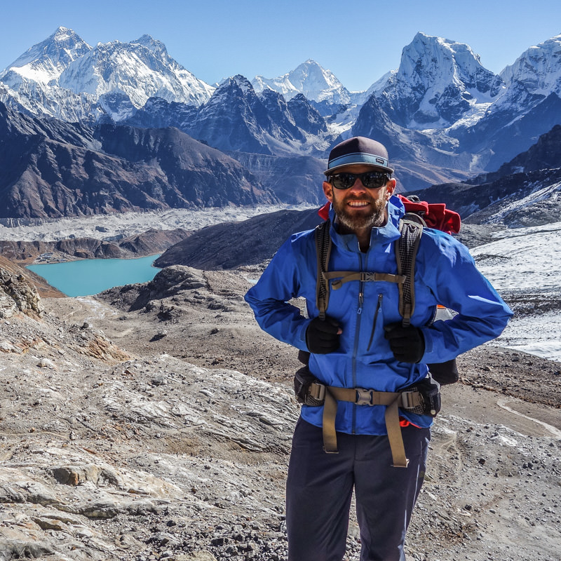 Dave Collins, a hiker standing in front of huge snowy mountain peaks with a big valley and a turquoise lake in the background in Nepal.