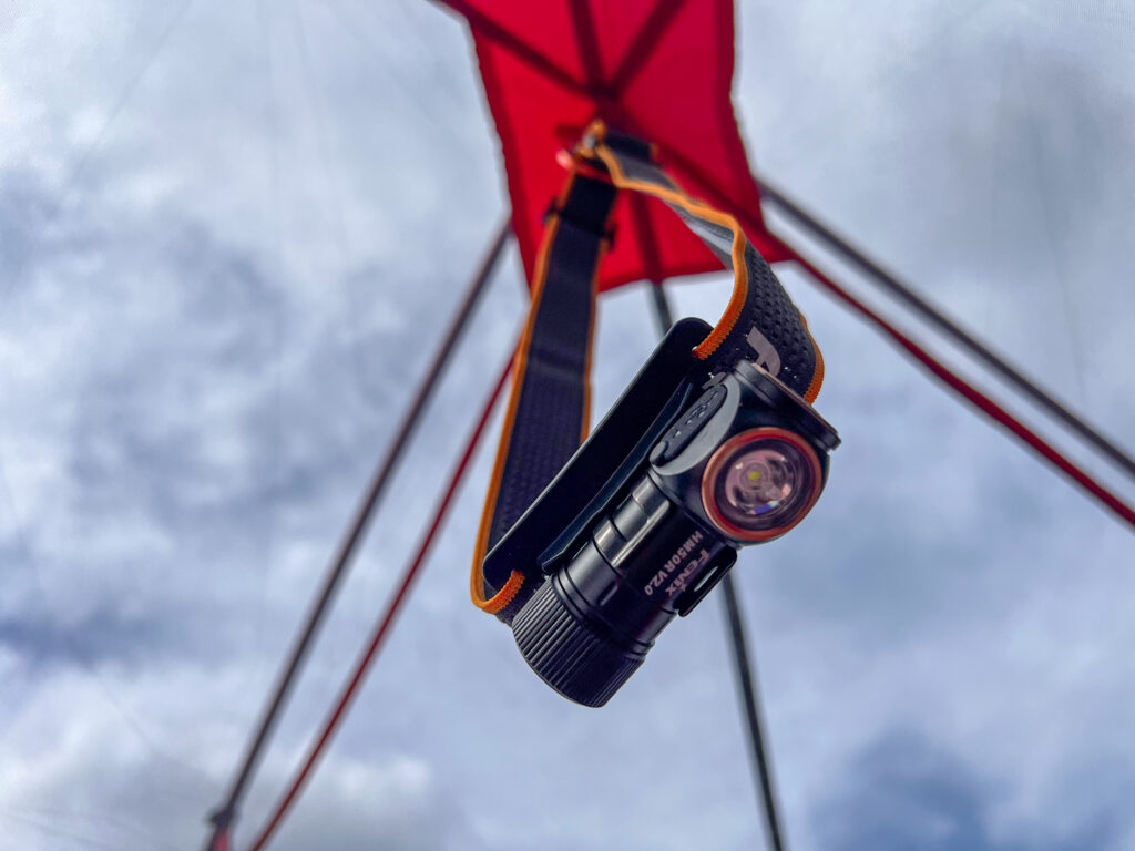 The image shows a Fenix HM50R V2.0 headlamp hanging upside down from a strap, with its beam pointed downward. The headlamp is positioned beneath a red tent fabric, and the sky is visible in the background.