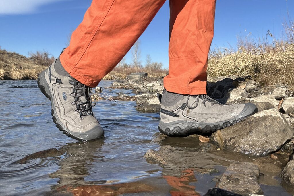 Close up image from knees down of a person wearing hiking boots and pants stepping out of a creek.