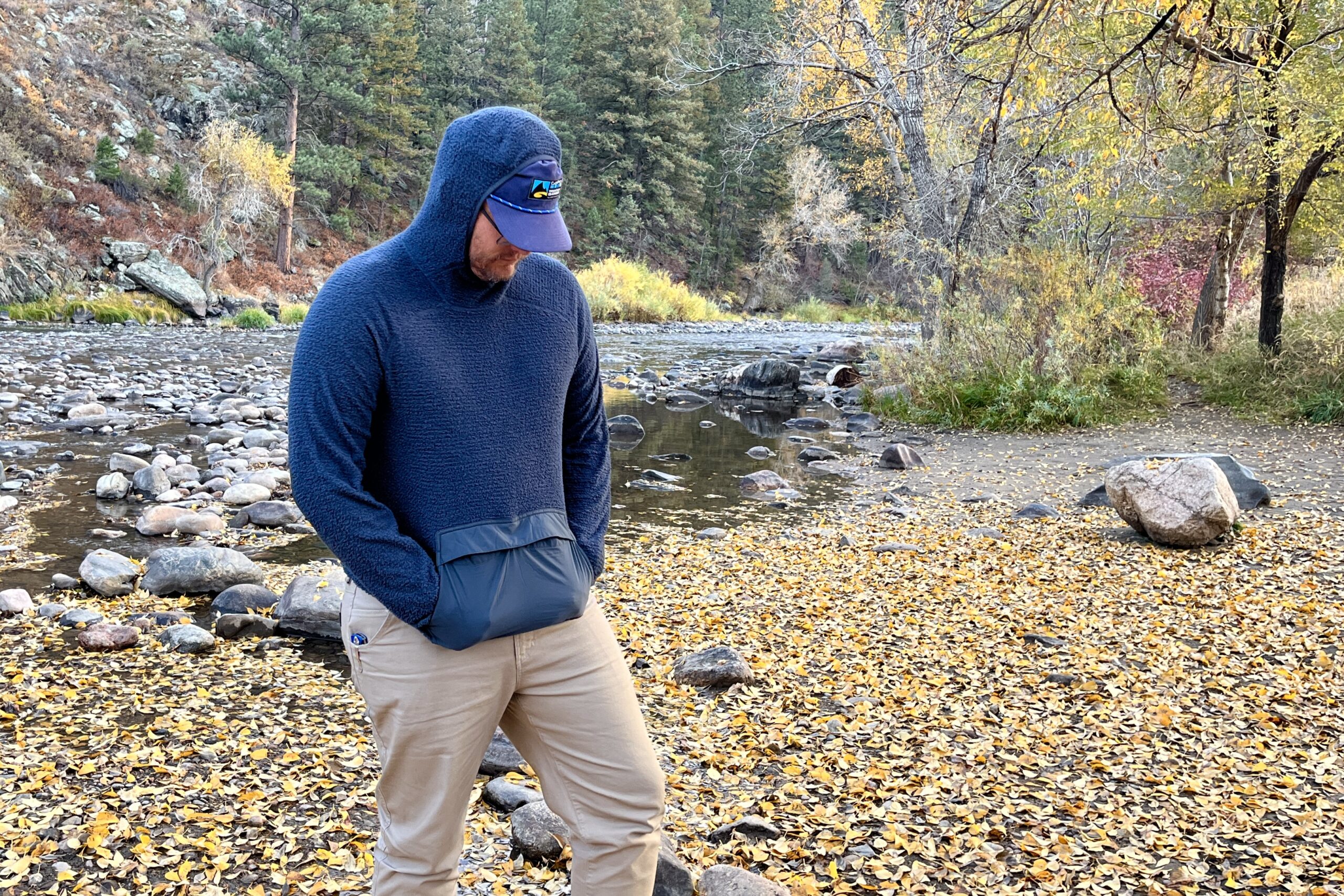 a man walking next to a river in the fall with an eddie bauer super sevens fleece on with the hood up and his hands in the kangaroo pouch pocket