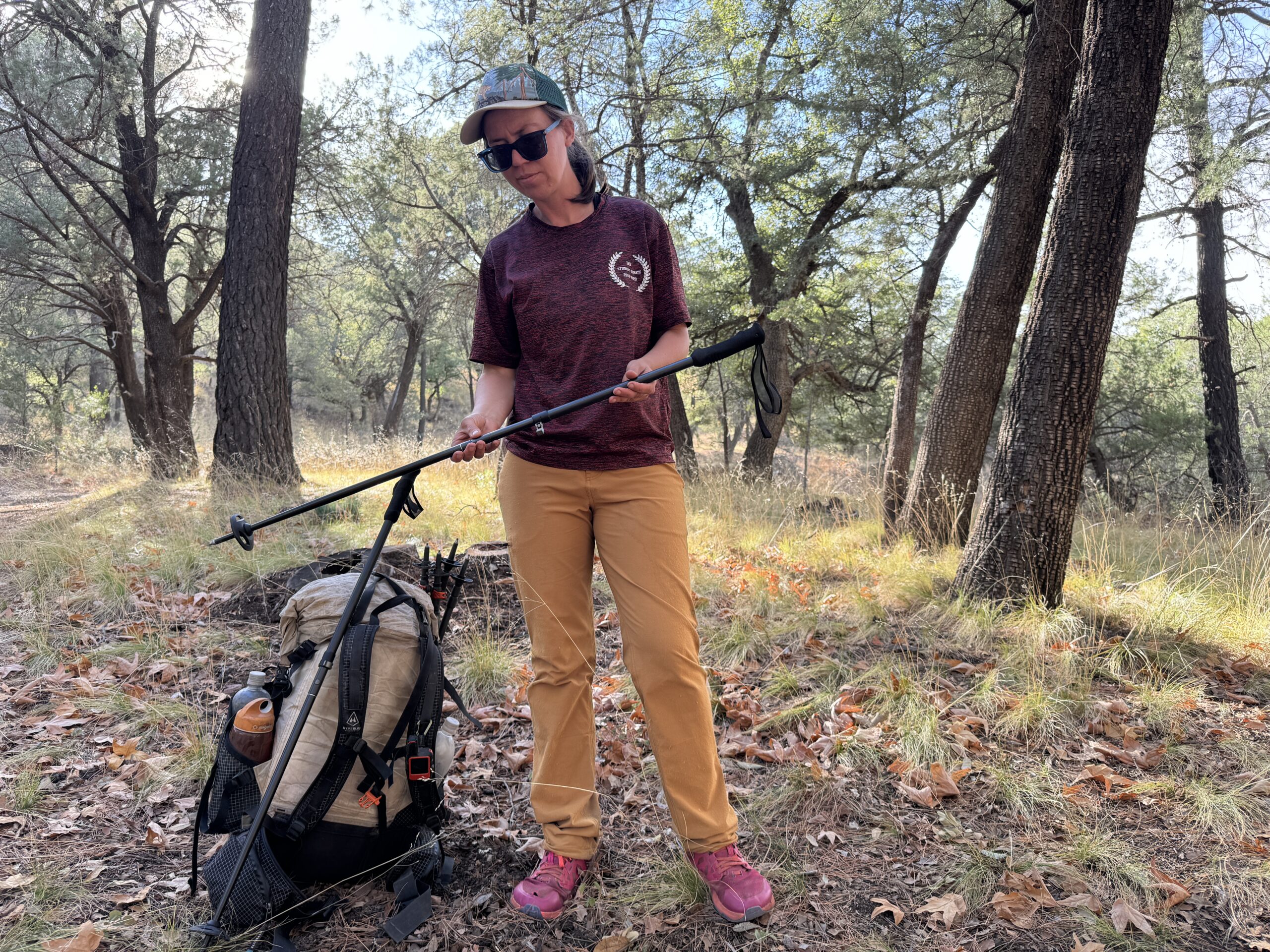 A hiker is extending a trekking pole while standing next to a backpack in the woods and wearing the Ferrosi pants.