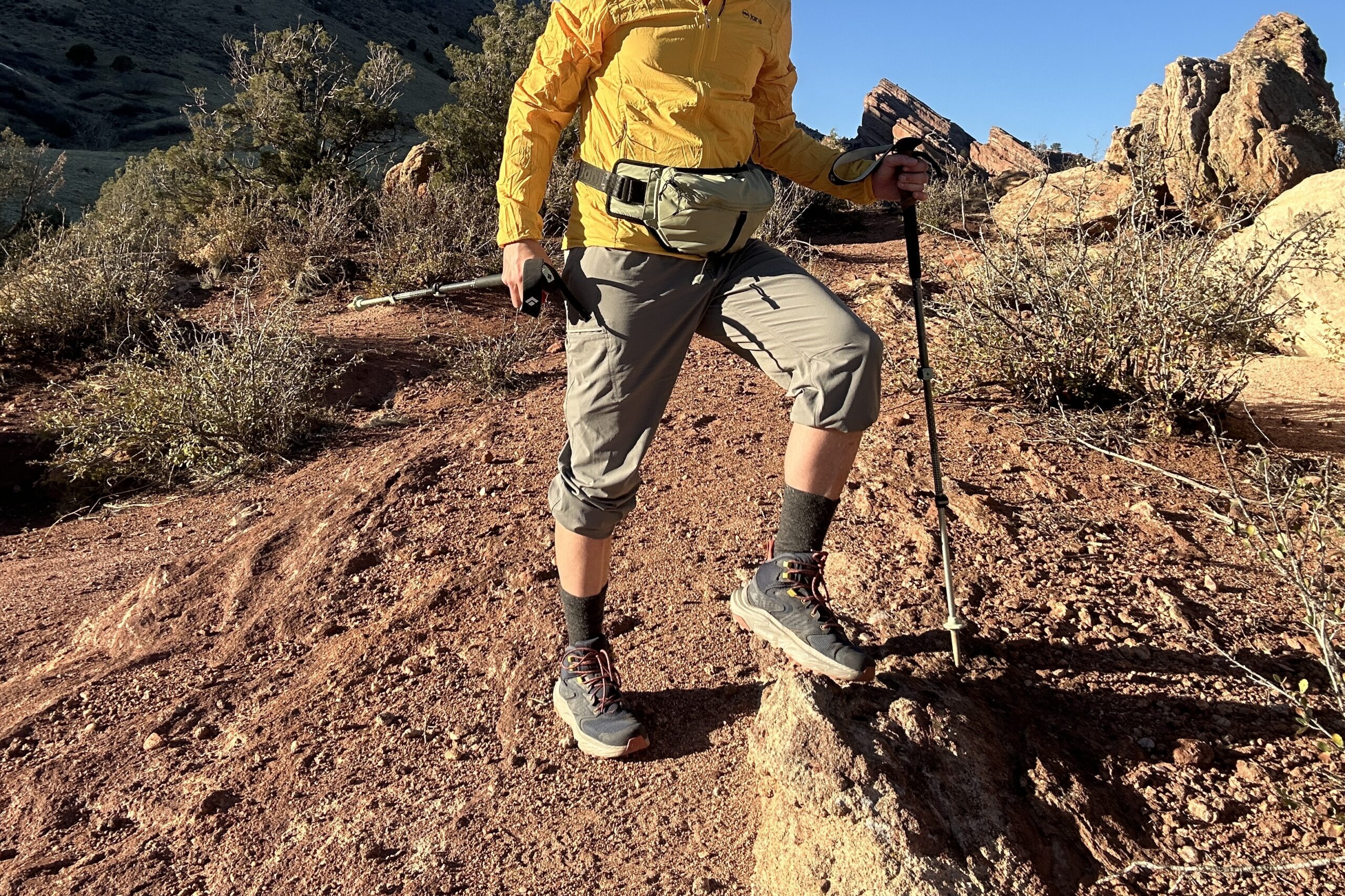 A man poses in a desert setting with boots on holding hiking poles.