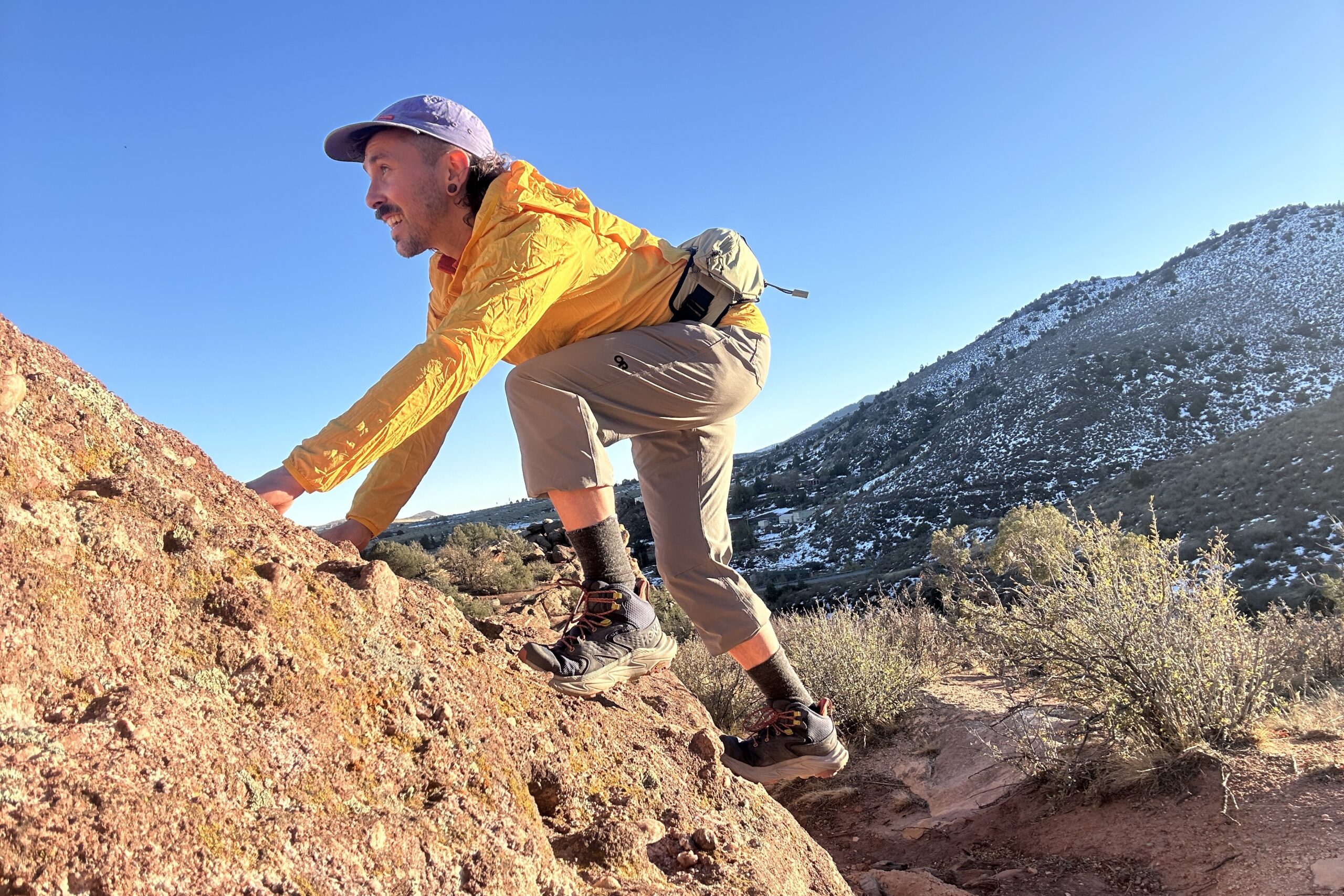 A man scrambles up some rocks in a cap and yellow jacket.