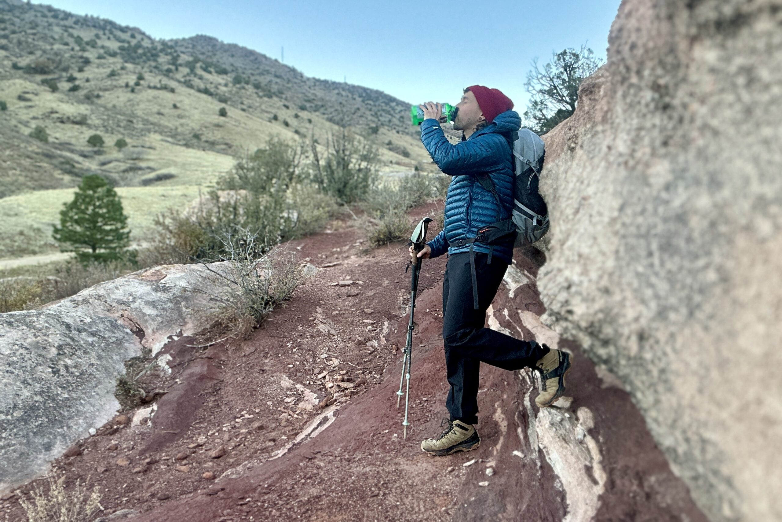 A man drinks out of a water bottle while leaning against a rock, wearinga hat and puffy and boots.