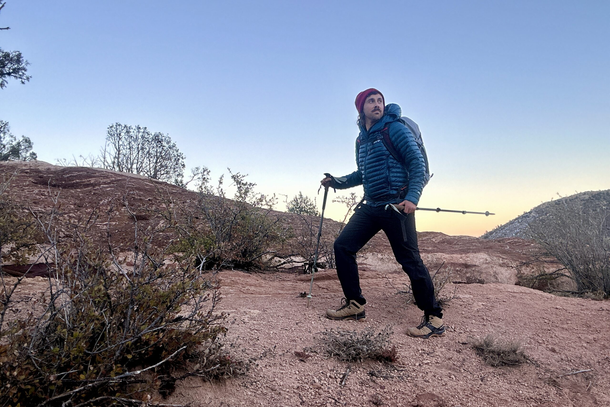 A man hikes in a puffy and beanie with a backpack and poles at sunset in a desert setting.