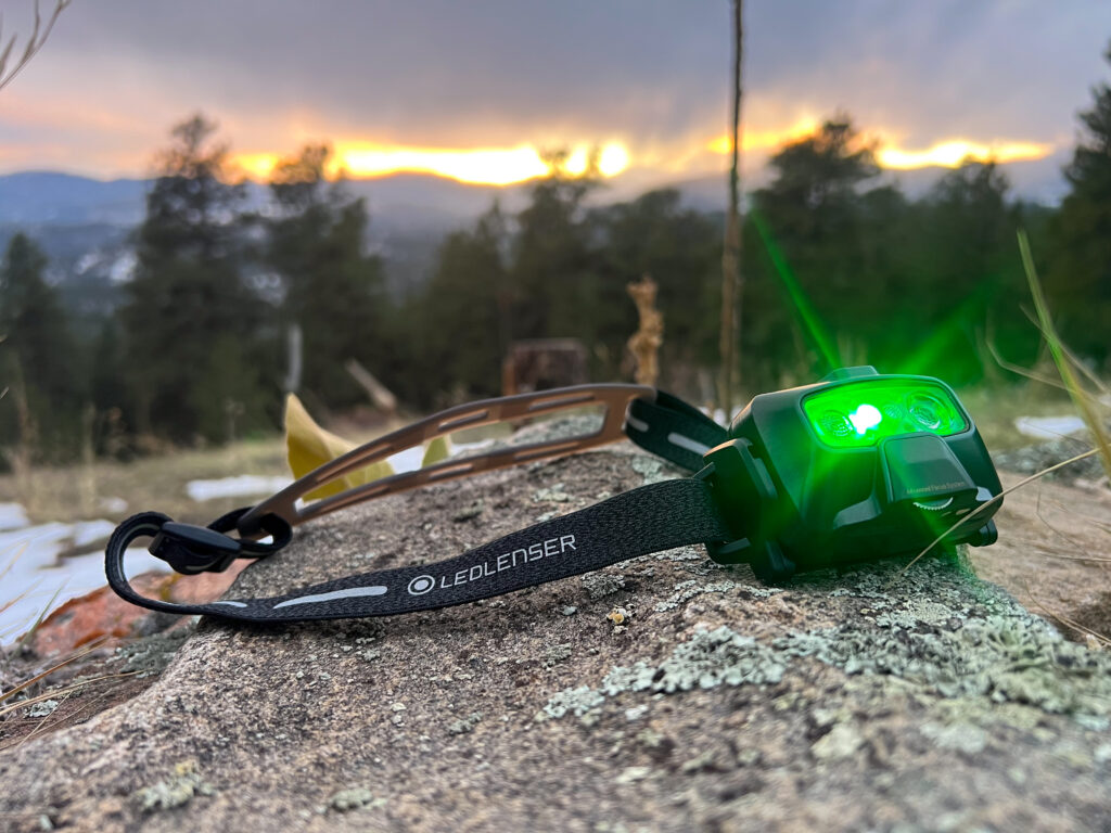 The image shows the Ledlenser HF6R Signature headlamp placed on a rock with a green light glowing from the front. The headlamp is resting on a natural surface with lichen, and the strap is partially visible. In the background, the fading light of a sunset creates a gradient in the sky, with trees and distant mountains framing the view.