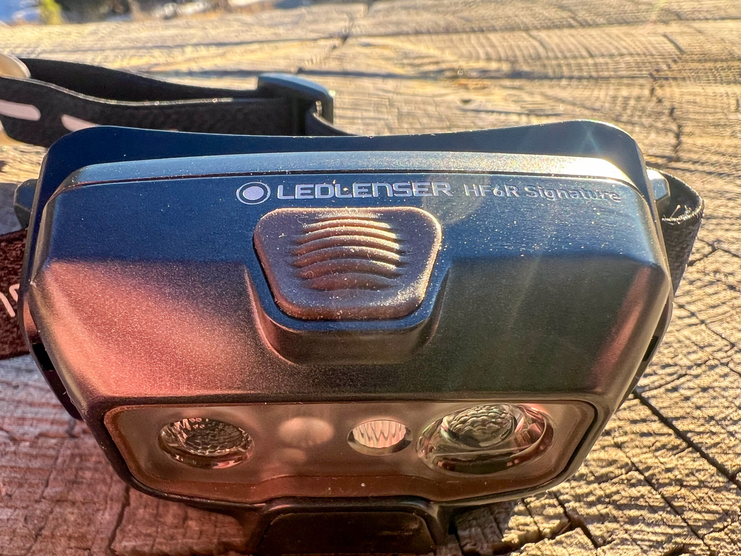 The image shows the front view of the Ledlenser HF6R Signature headlamp, focusing on the power button located at the top. The headlamp is resting on a wooden surface with sunlight reflecting off its body.