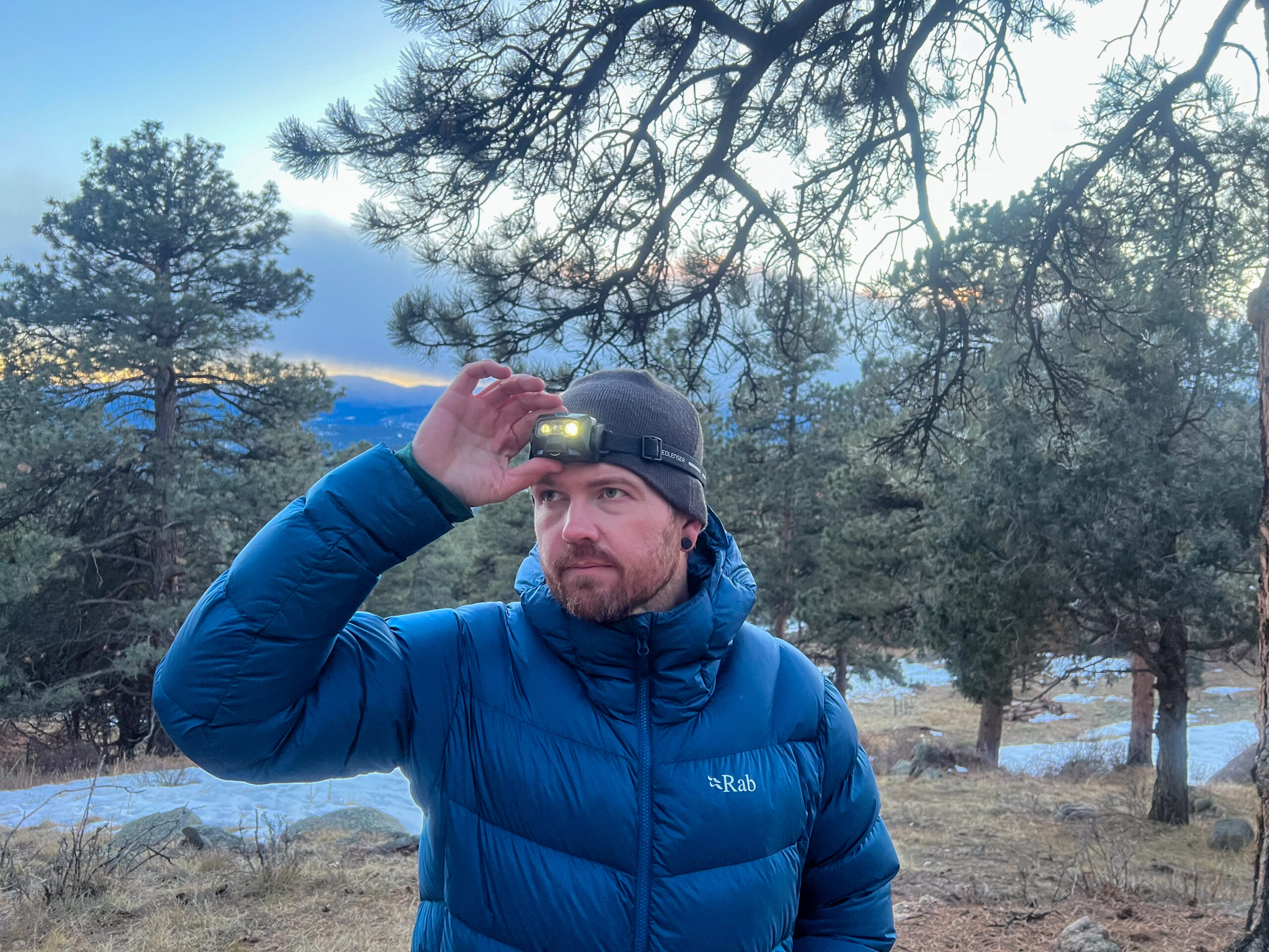 The image shows a person wearing a blue down jacket and a beanie while adjusting a Ledlenser HF6R Signature headlamp on their forehead. The background features trees with a sunset sky and snow-covered ground.