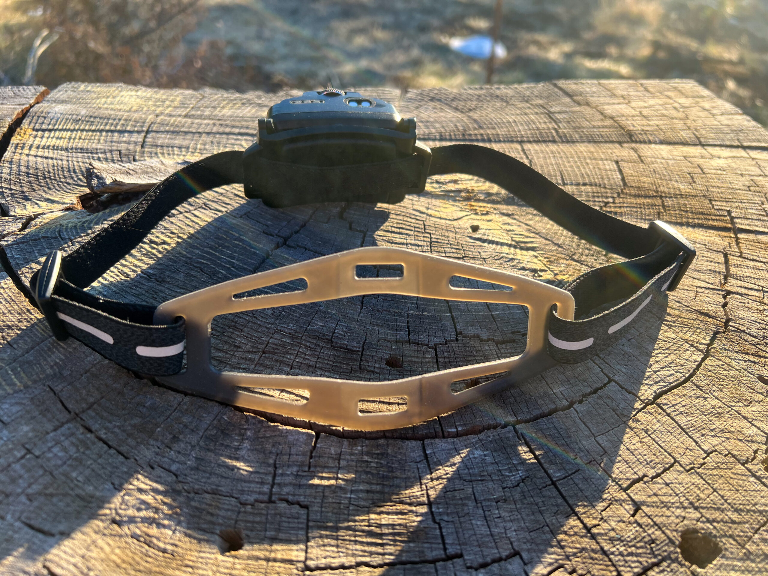 The image shows the Ledlenser HF6R Signature headlamp strap laid across a weathered wooden surface. The strap is black with reflective stripes. The sunlight creates a warm glow in the background, highlighting the contours of the strap and the texture of the wood beneath.