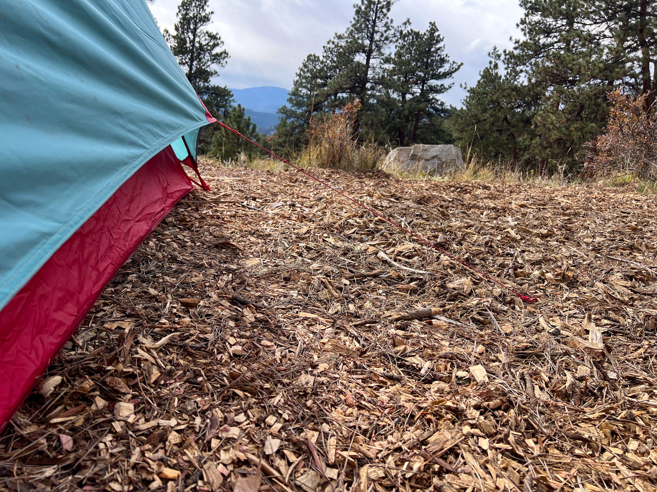 A close-up view of the base of the tent, showing the pink and teal fabric with a guyline staked into the ground. The ground is covered in wood chips, and the background shows a forested area with trees and distant rocks.