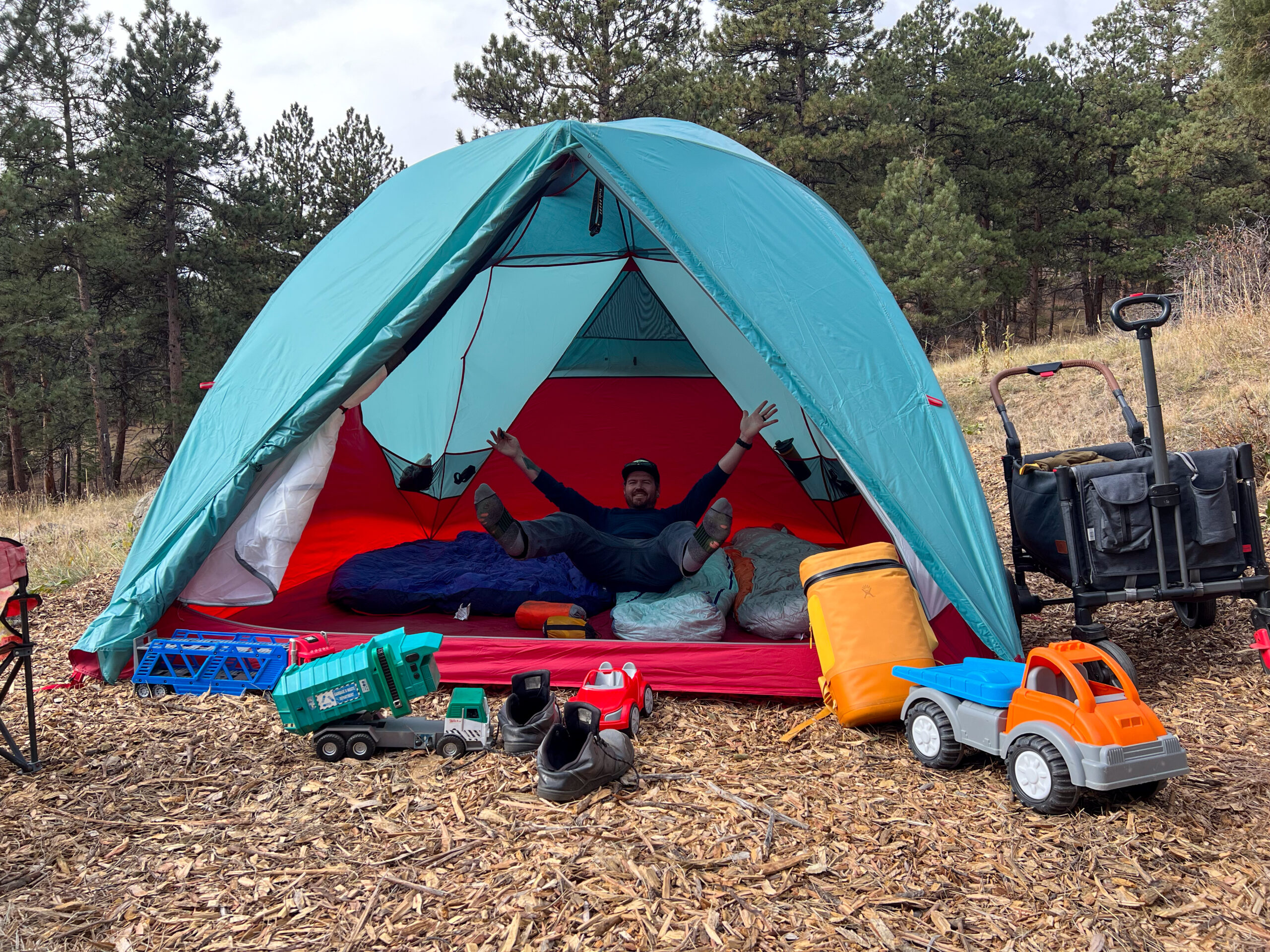 A person is sitting inside the spacious MSR Habitude 6 tent, surrounded by sleeping bags, children’s toys, and camping gear. The large interior is well-lit, with plenty of room for gear and sleeping arrangements.