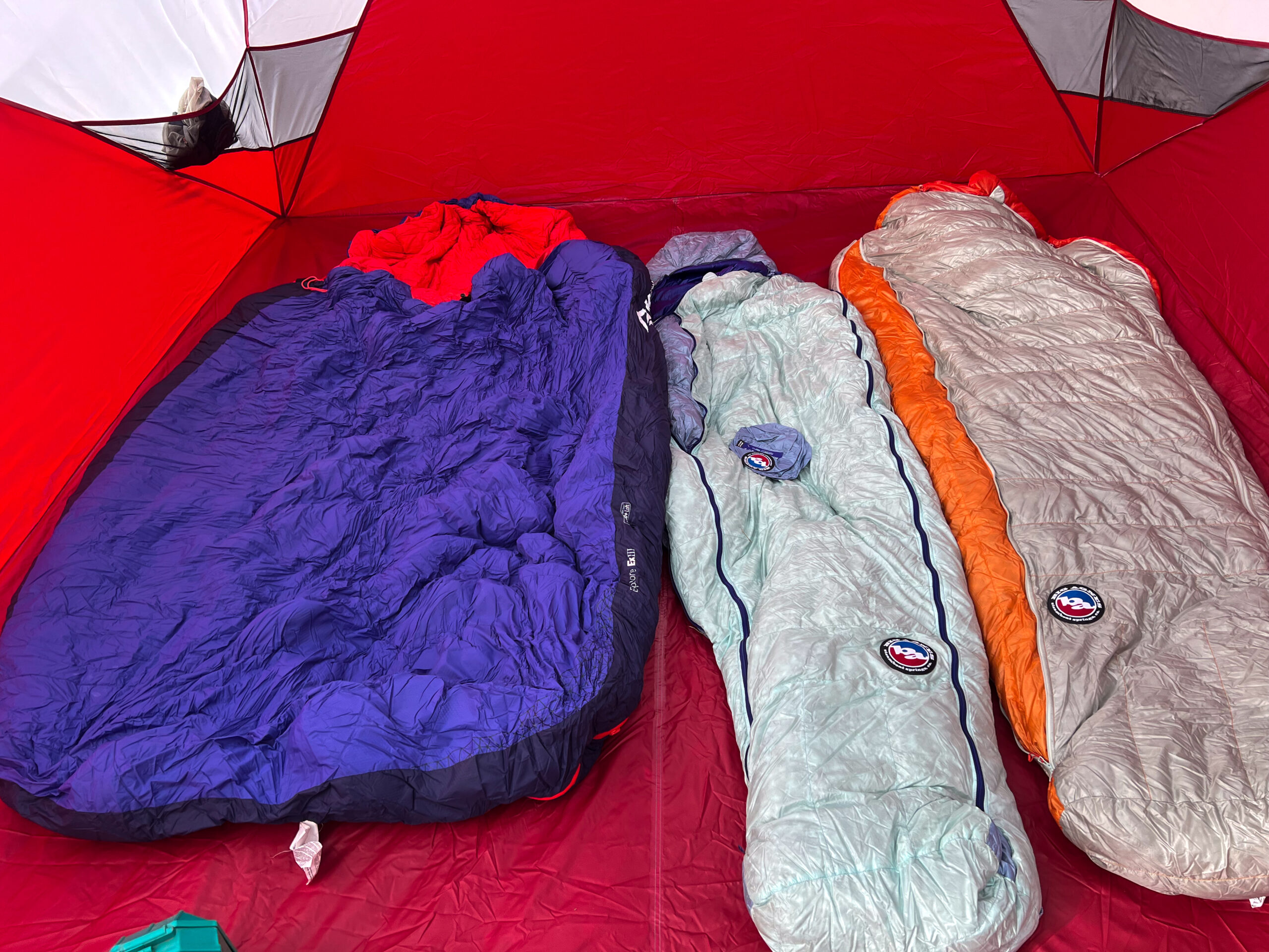 Inside the MSR Habitude 6 tent, three sleeping bags are laid out on the red tent floor. The bags are arranged side by side with different colors: purple, greenish-blue, and light gray/orange. The spacious interior showcases plenty of space for family camping.