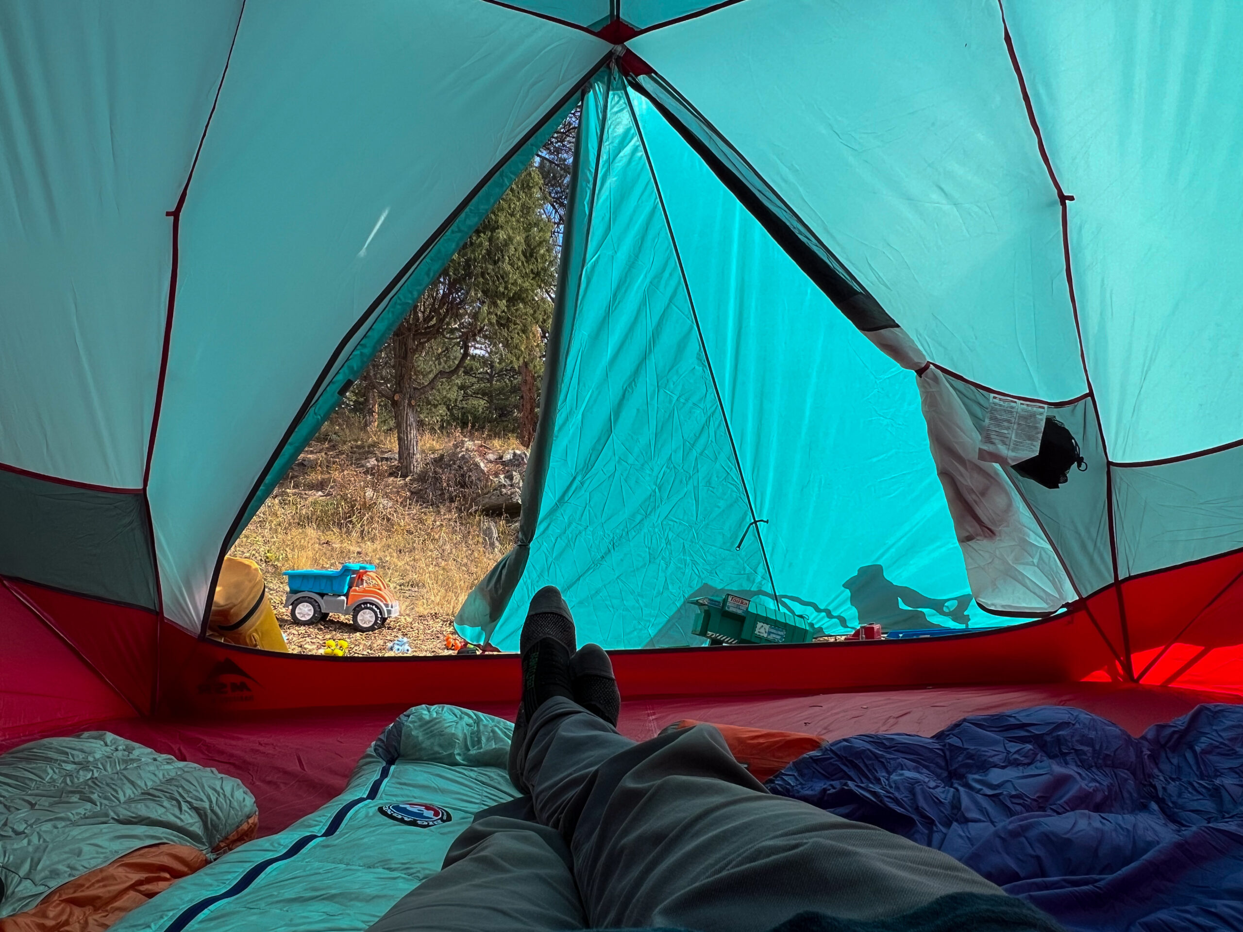 View from inside the MSR Habitude 6 tent showing a person reclining with their legs stretched out in front. In the background, a toy truck is visible outside through the large, bright turquoise tent windows. The interior features sleeping bags and plenty of space for relaxation.