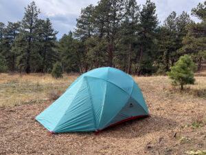 A side view of the MSR Habitude 6 tent set up in a forested area, showcasing the colorful rainfly and its dome-like shape, with tall trees in the background.