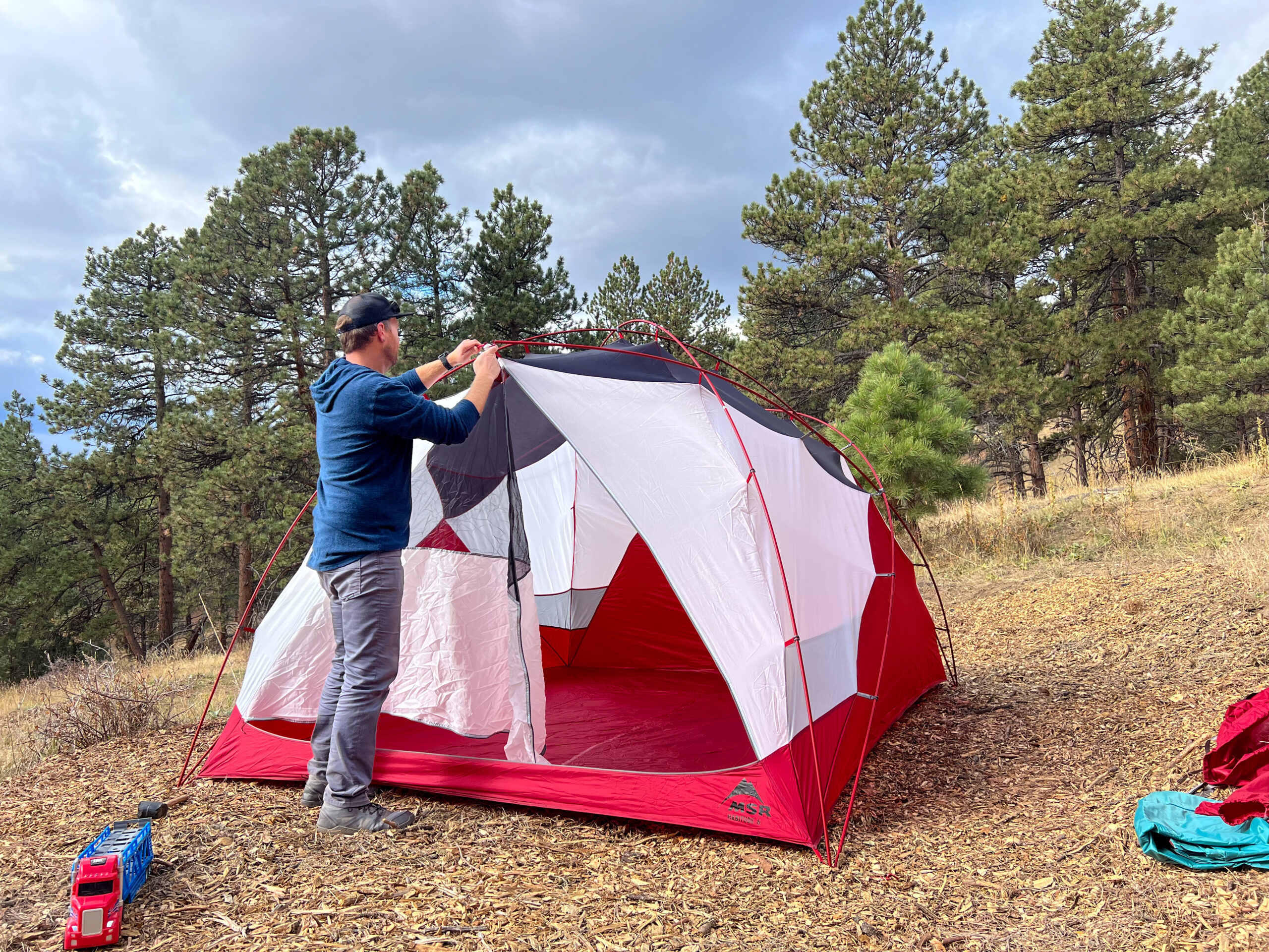 A person is setting up a red and white tent in a forested area. They are attaching the mesh door to the frame, and a toy truck is placed nearby on the ground. The tent is large, with a spacious interior and a wooded backdrop.