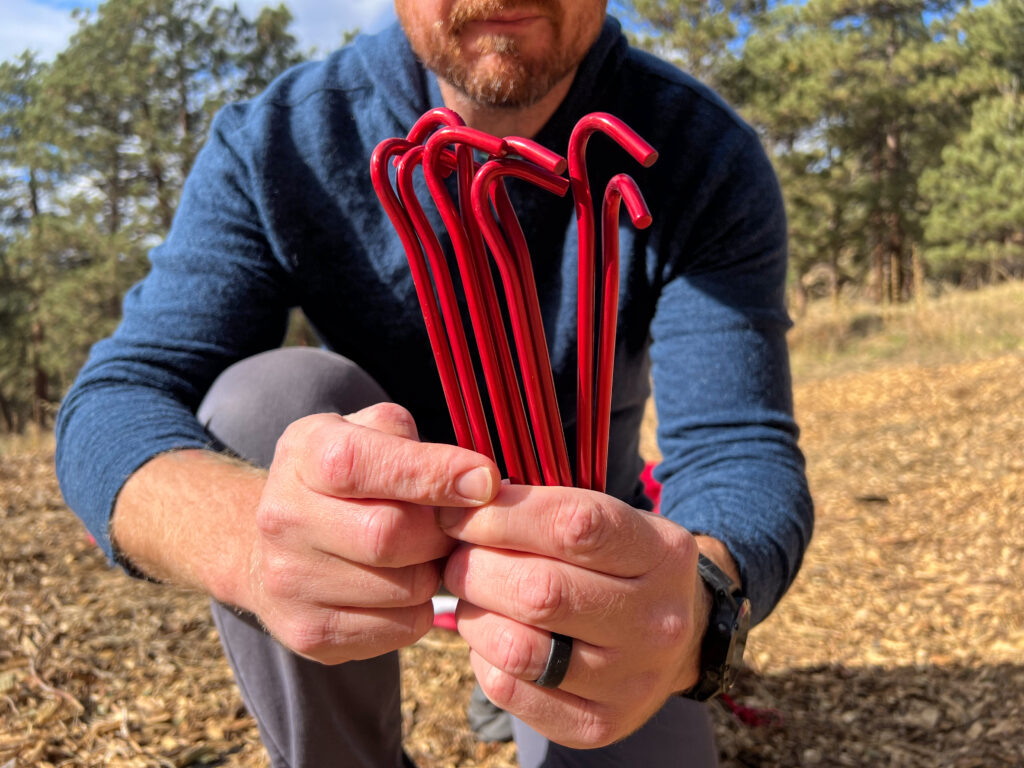 A person holding several red tent stakes in their hands, with the stakes visibly bent in a "hook" shape. The background shows a wooded area with scattered dry leaves.