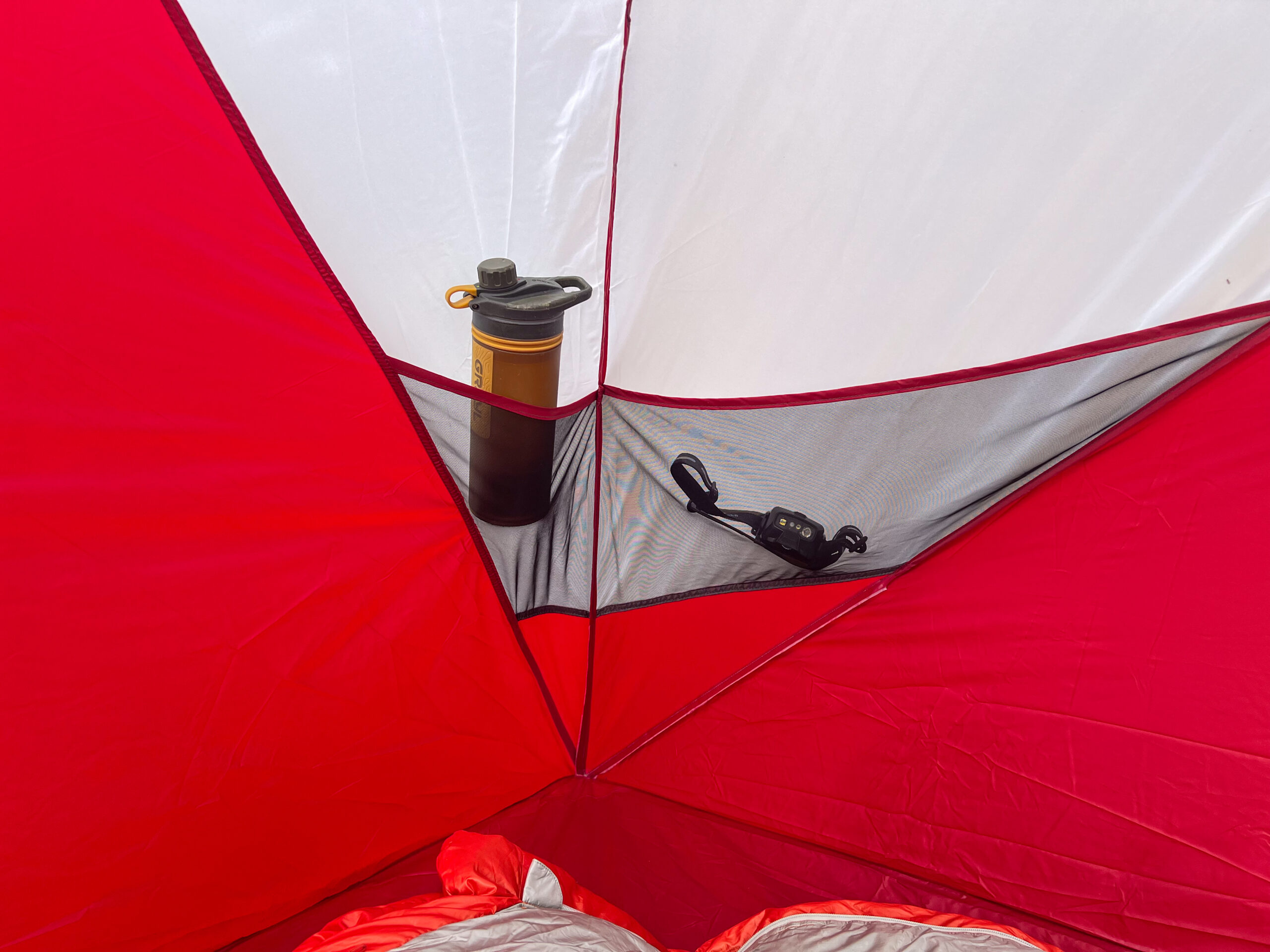 The interior of the MSR Habitude 6 tent showing a mesh storage pocket holding a water bottle and a small headlamp. The red and white tent fabric is visible, with a sleeping bag partially shown in the foreground.