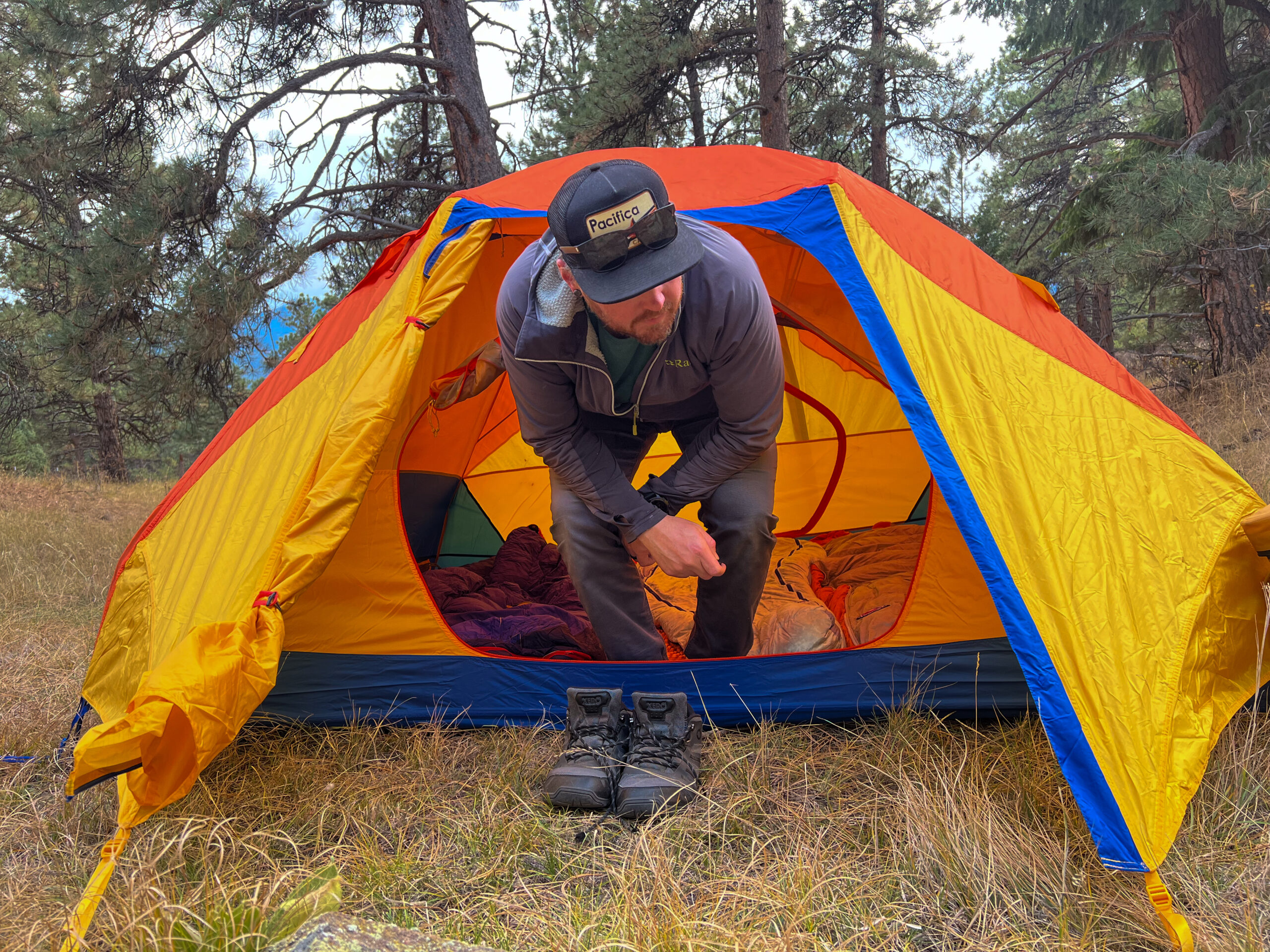 A camper crouches while exiting a brightly colored tent, highlighting the low peak height of the tent. The vibrant orange, yellow, and blue tent is pitched in a wooded campsite surrounded by trees. A pair of hiking boots is positioned outside the tent on the grassy ground.