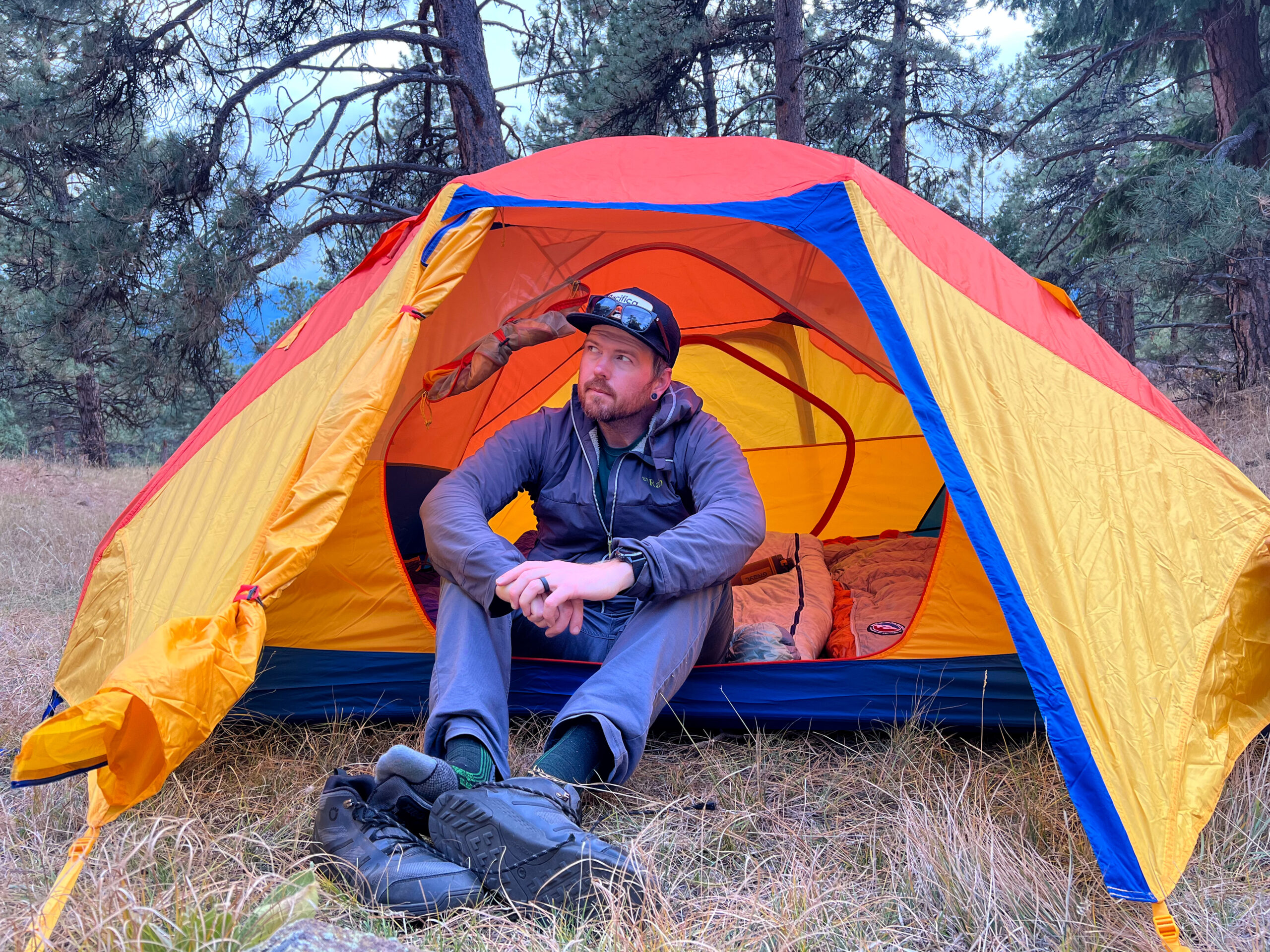 A camper sits cross-legged in the entrance of a brightly colored tent, showcasing its low headroom and cozy interior. The vibrant orange, yellow, and blue tent is pitched in a wooded area, with a pair of hiking boots placed on the grassy ground outside.