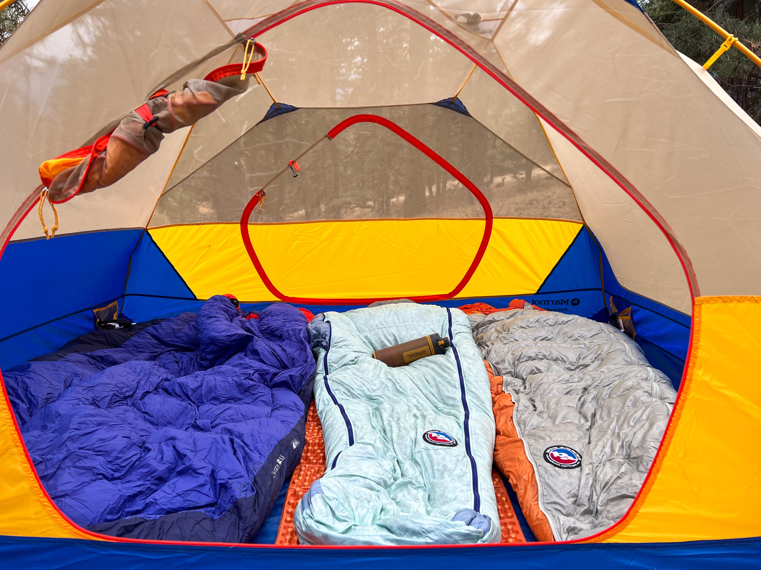 Interior view of the Marmot Tungsten 4p tent with three sleeping bags and camping gear neatly arranged, showcasing its spacious layout and vibrant color scheme at a forest campsite.