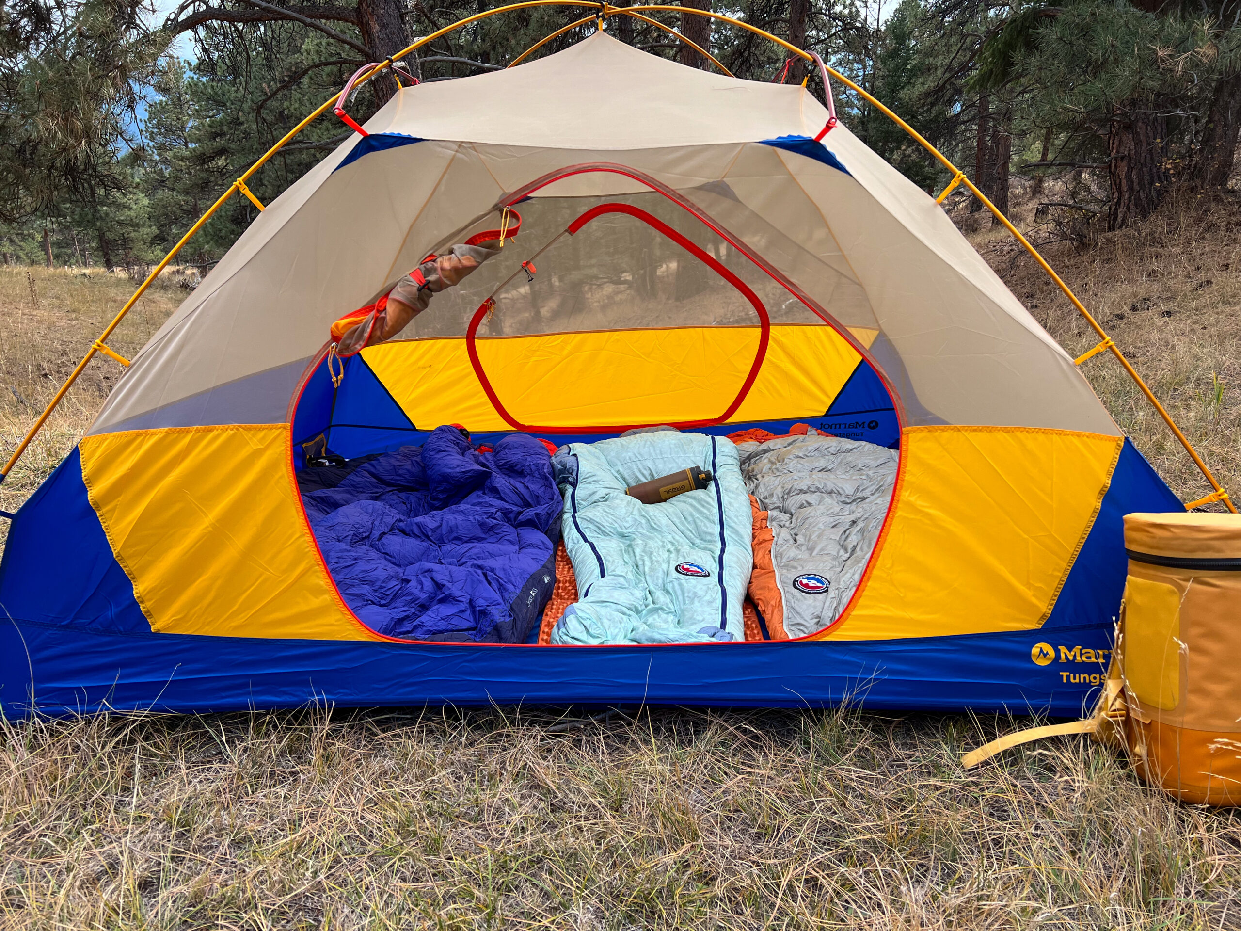 Marmot Tungsten 4p tent set up in a grassy campsite, showcasing its spacious interior with three sleeping bags and a cooler placed outside the tent.