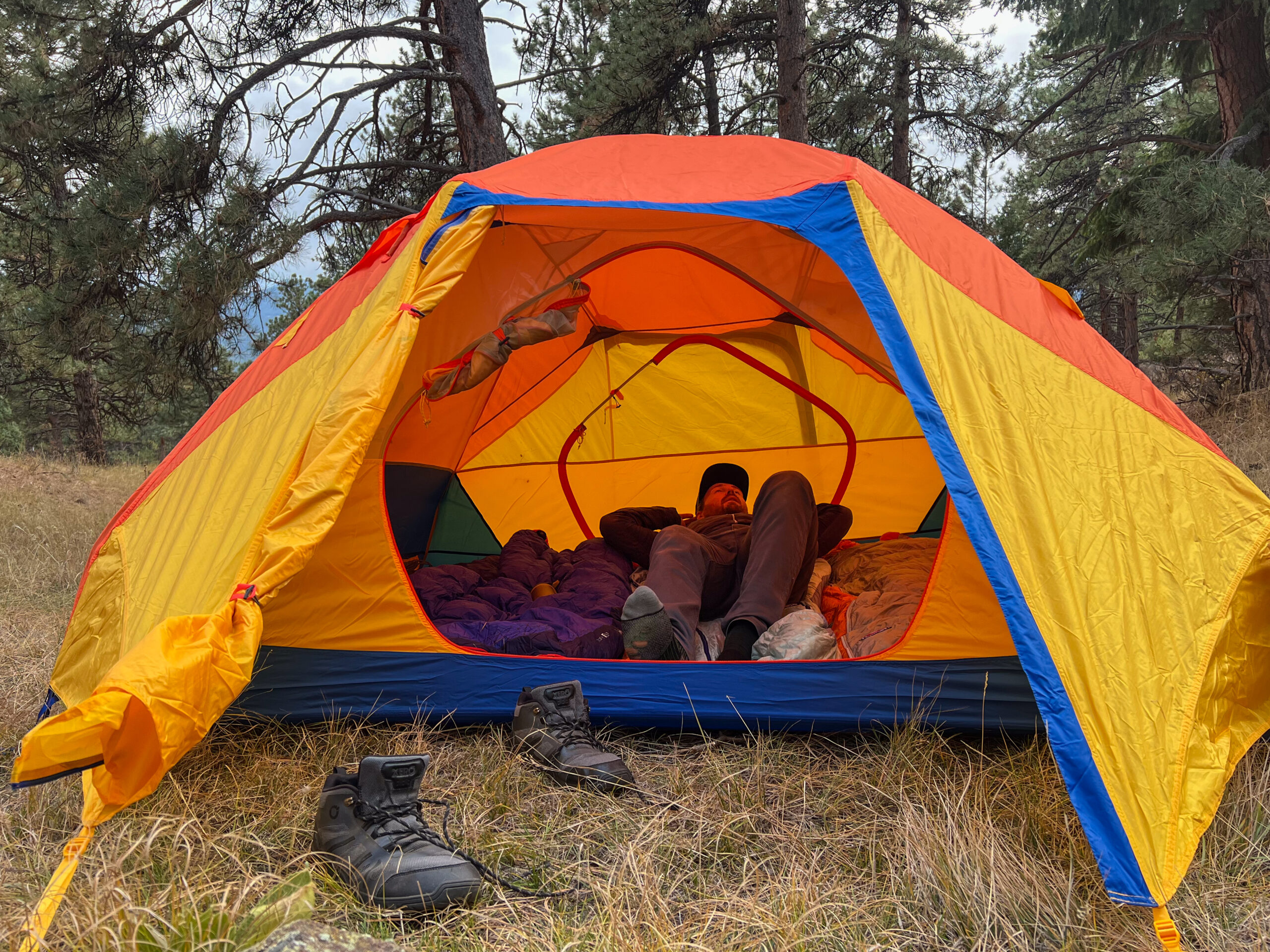 Camper lounging inside a spacious Marmot Tungsten 4p tent with its vibrant red and yellow exterior, set up in a grassy forest clearing, with boots placed just outside the entrance.