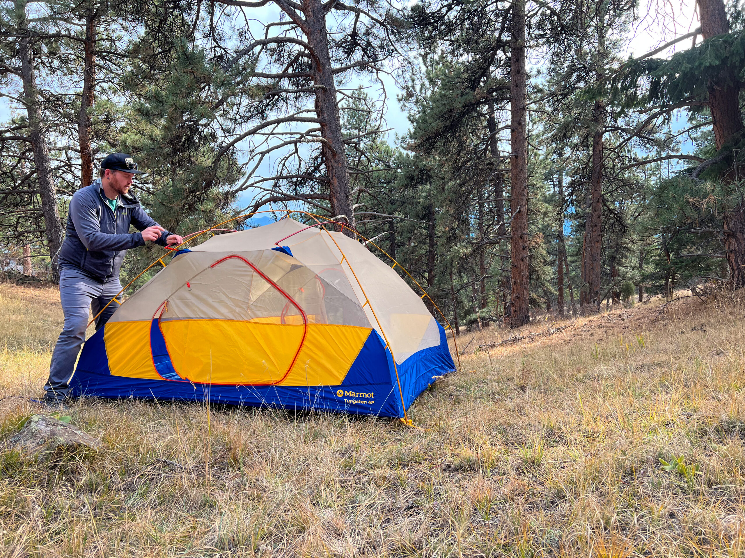 A camper setting up the Marmot Tungsten 4p tent in a wooded campsite, showcasing the tent's easy assembly with its color-coded pole system and spacious design.