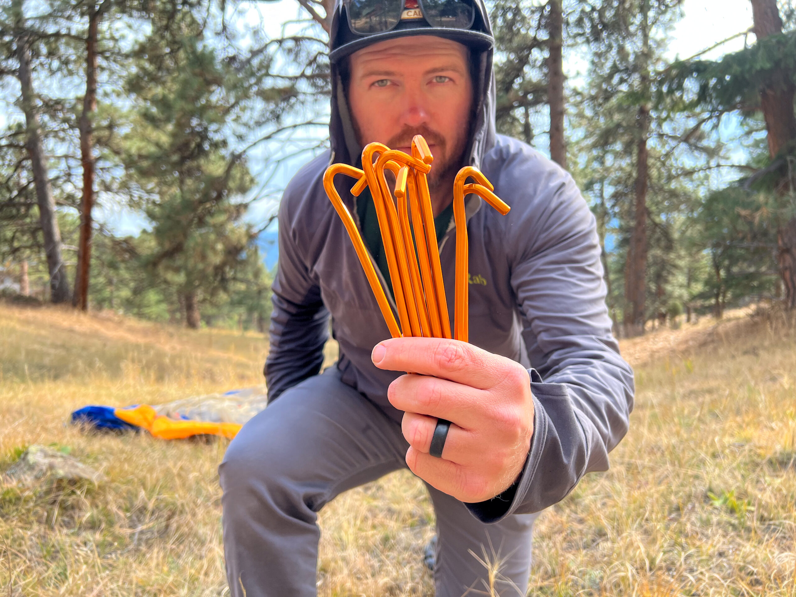 A person holding a handful of bright orange tent stakes in front of them, with a forested background and the Marmot Tungsten 4 tent in the distance.