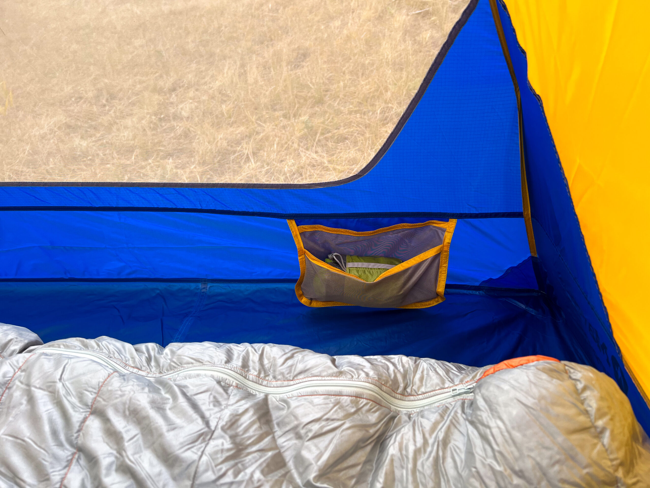 A close-up view of the interior of the Marmot Tungsten 4 tent, showing a mesh storage pocket on the sidewall with some gear inside, alongside a sleeping bag. The tent features a colorful blue and yellow design.