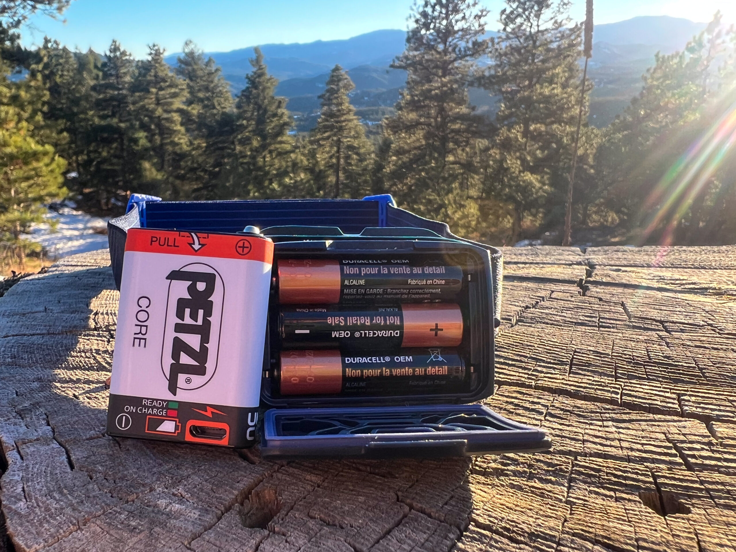 The image shows the battery compartment of a Petzl Actik headlamp with a visible comparison between the Petzl CORE rechargeable battery and disposable AAA batteries. he headlamp's open battery compartment is set on a wooden surface with a scenic outdoor background.