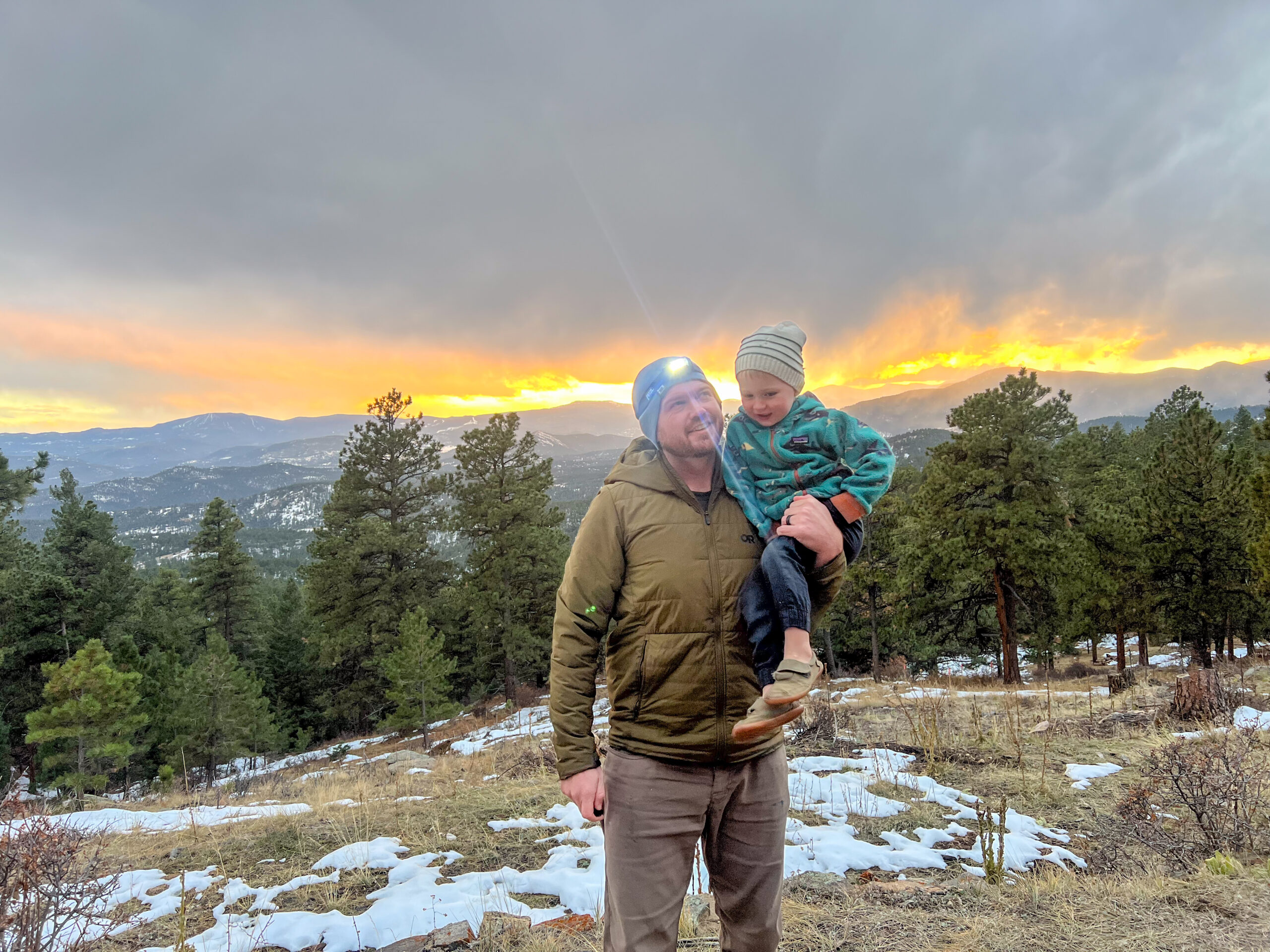 The image features a father holding his young son with the sun setting in the background. Both are wearing warm outdoor clothing. The father is wearing a Petzl Actik headlamp, and both are smiling as they look at the camera.