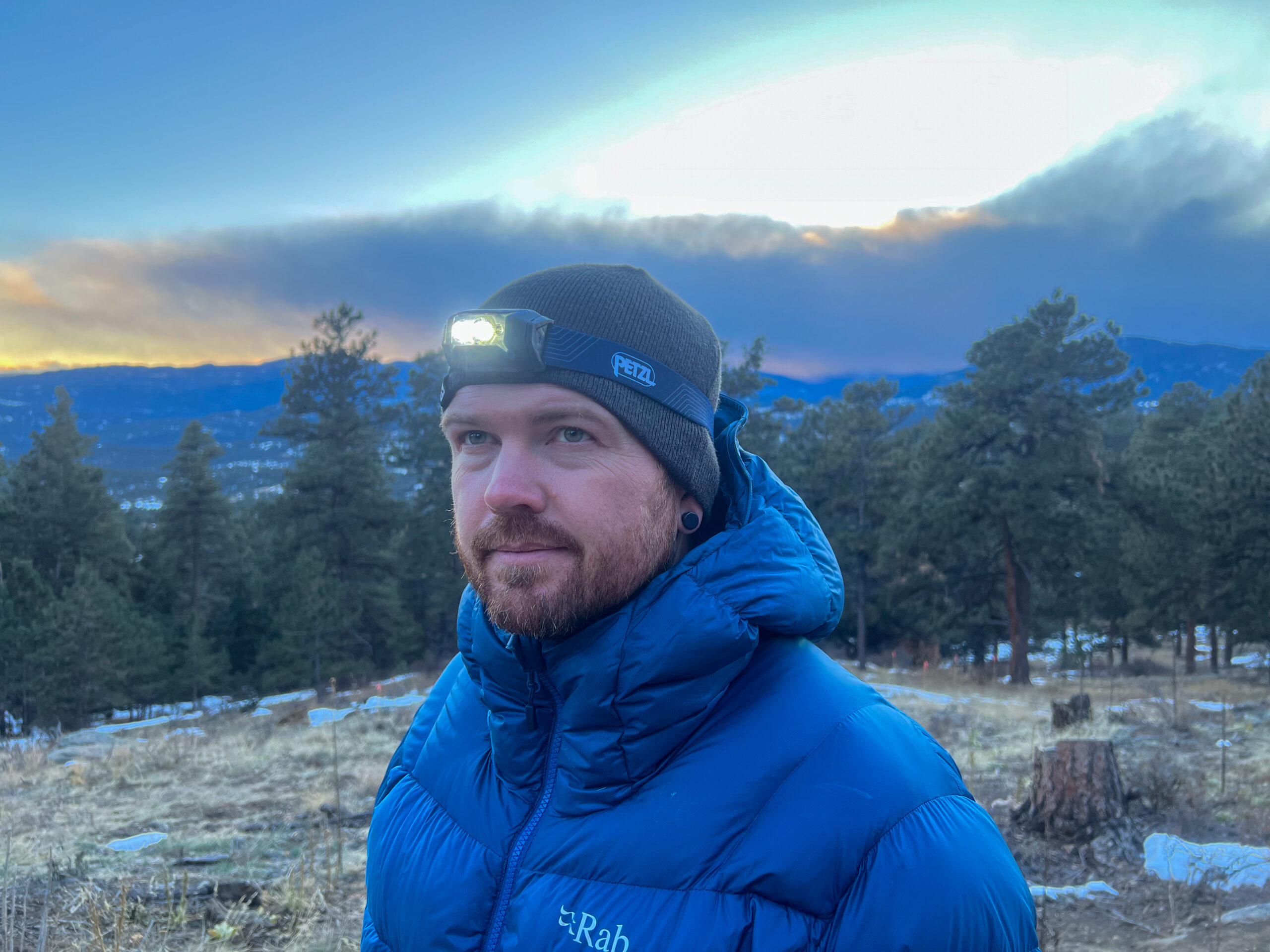 The image shows a man wearing a Petzl Actik headlamp, standing outdoors during dusk. He is dressed in a blue puffer jacket and a black beanie, looking off to the side. The background features a view of trees and mountains with the sky transitioning to evening. The headlamp is lit, casting light into the environment.