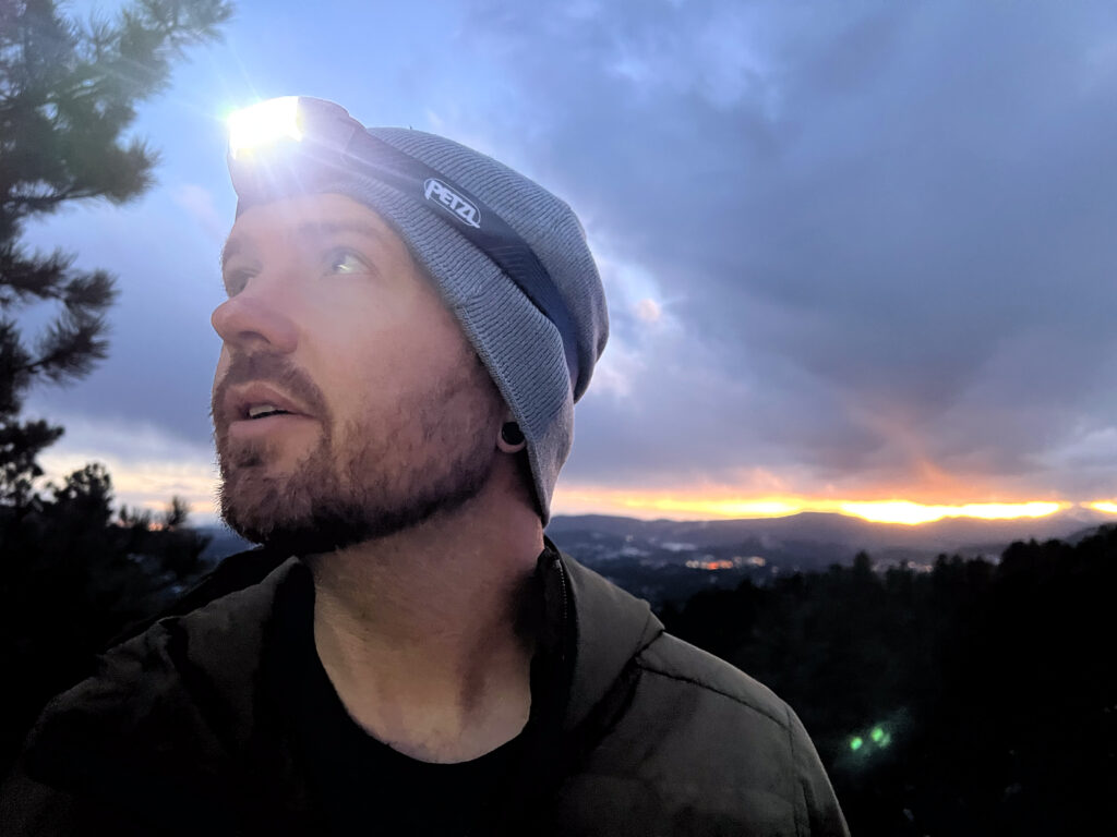 The image shows a person wearing a Petzl Actik headlamp while standing outdoors during dusk. The person is looking upward, with the headlamp illuminated, casting light into the surrounding environment. The sun is setting in the background