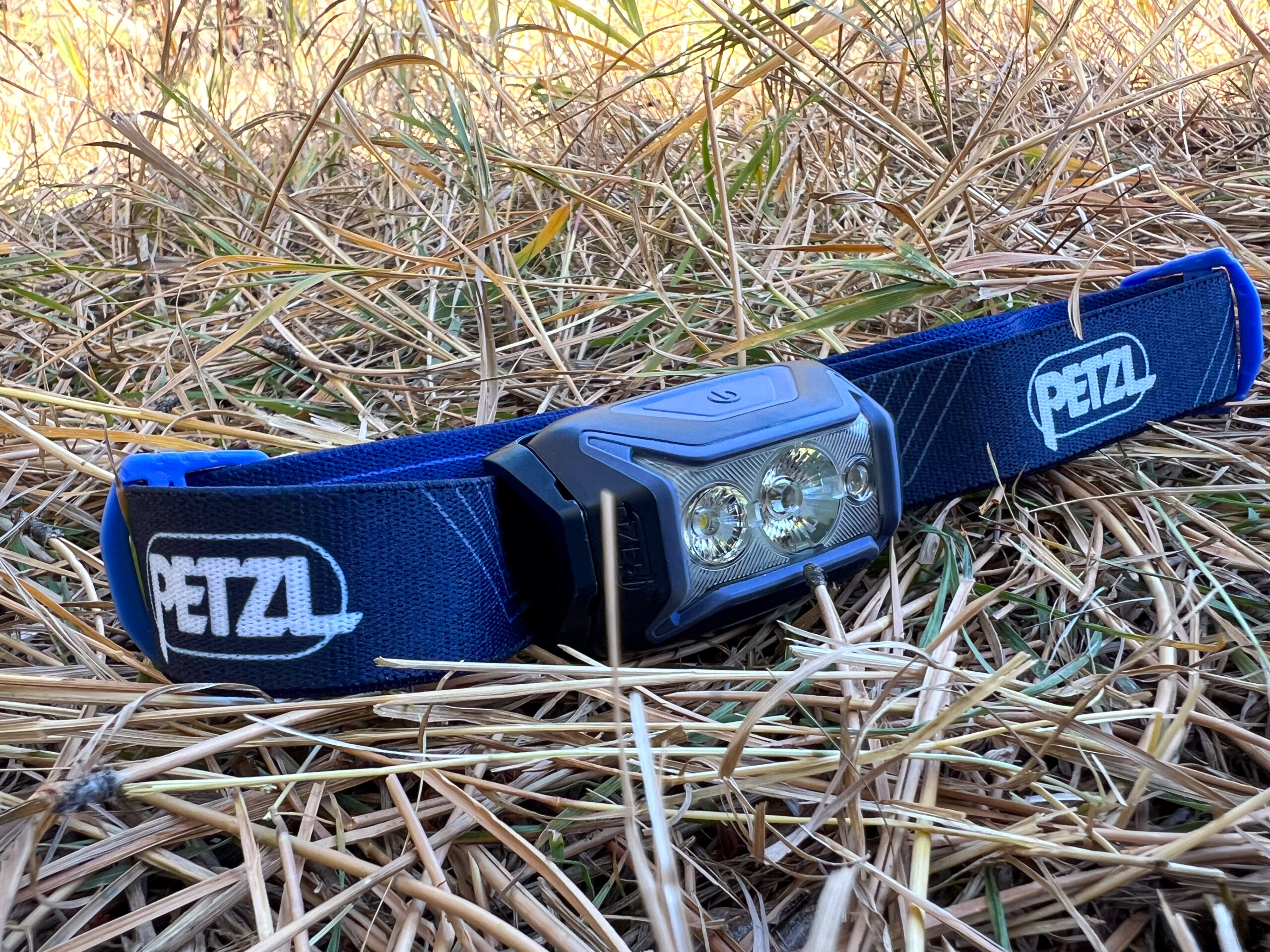 The image shows a Petzl Actik headlamp with a blue and black strap resting on dry grass.