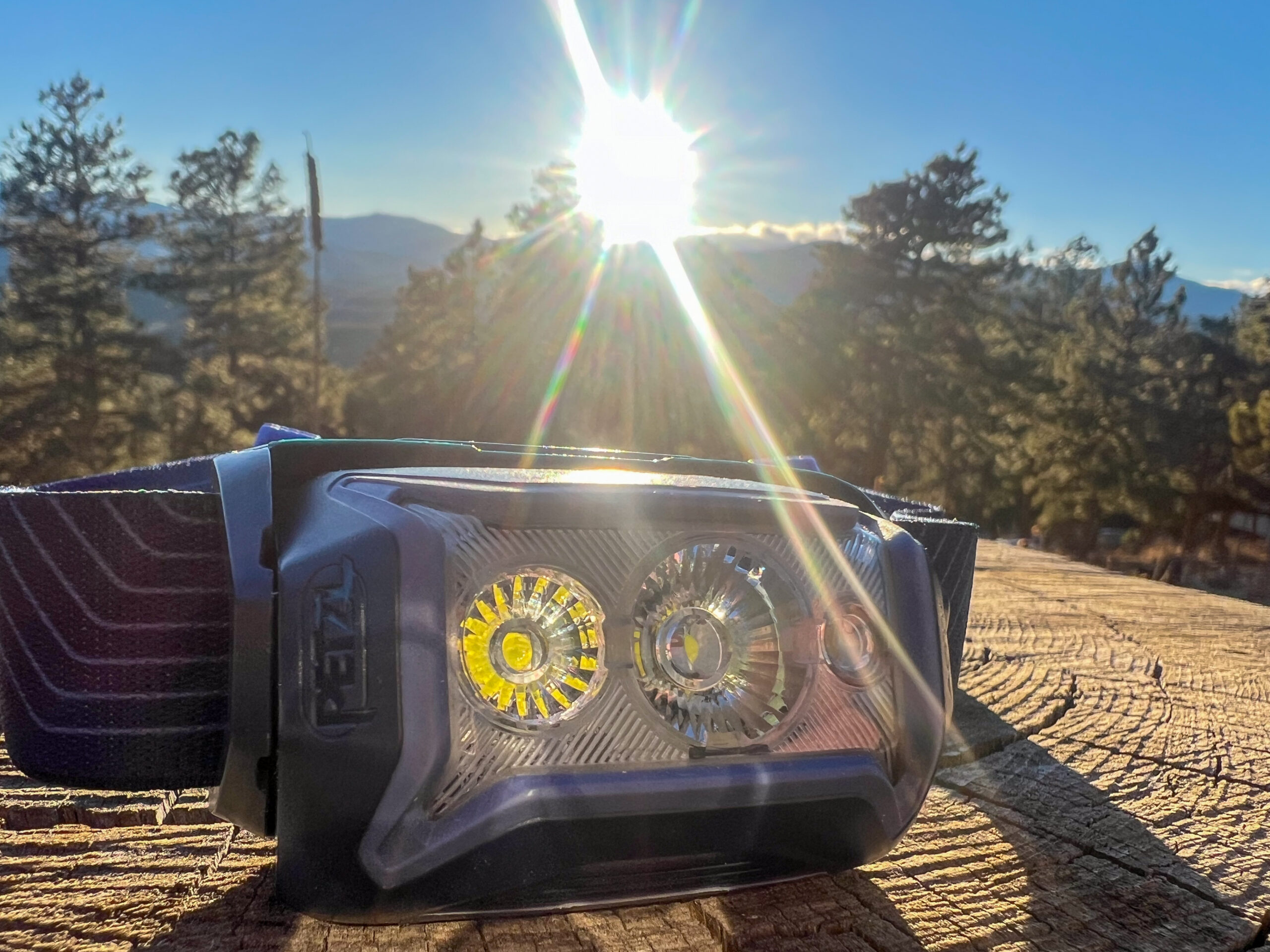 The image shows a close-up of the Petzl Actik headlamp with the sun in the background, creating a lens flare effect. The headlamp's lens features two large LED bulbs, one surrounded by a reflective ring. The headlamp is mounted on a black and blue strap, and the background consists of a mountainous landscape with pine trees and a bright, clear sky.