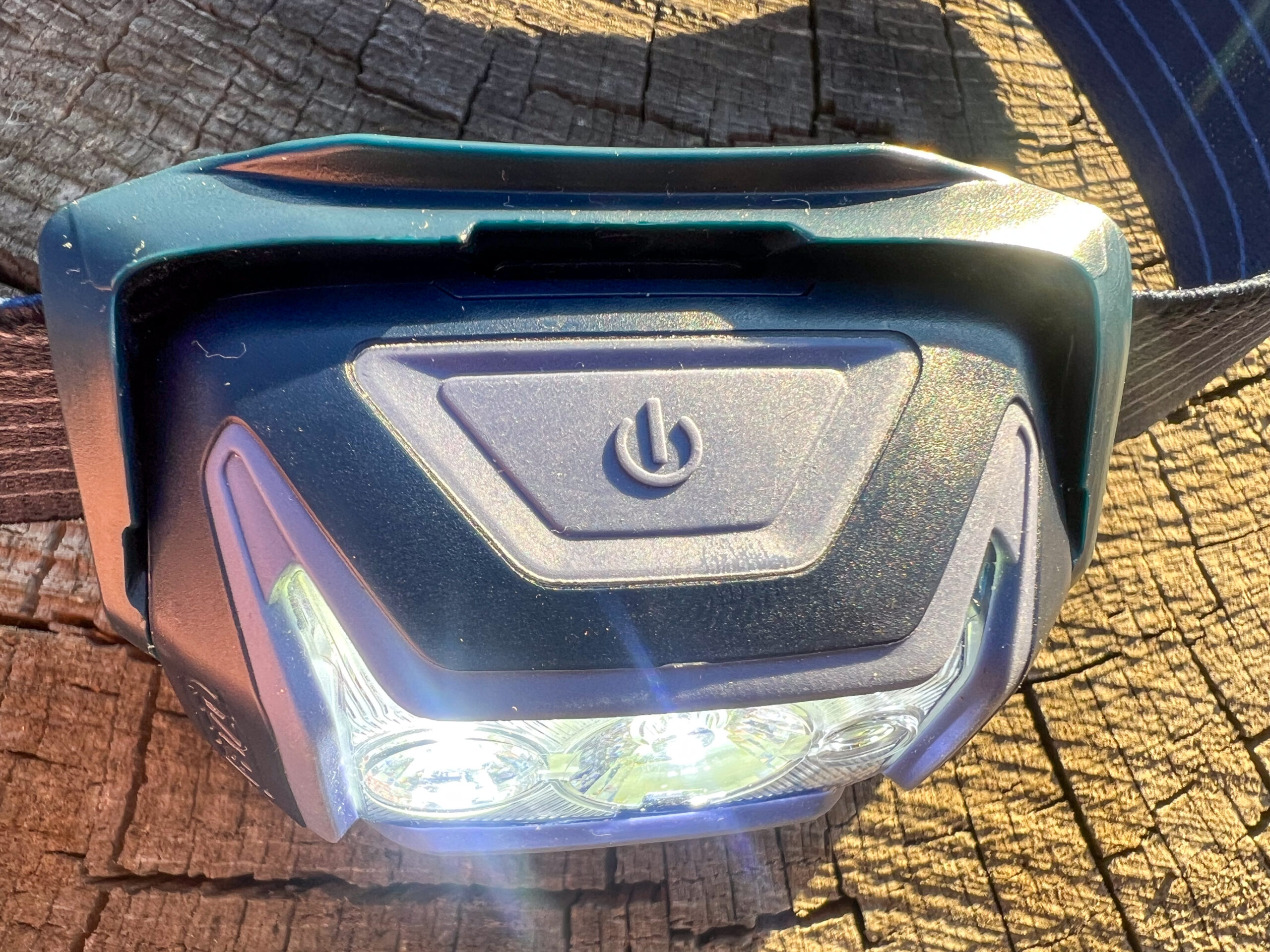 The image shows a close-up of the Petzl Actik headlamp's power button. The headlamp is positioned on a wooden surface, with the sun reflecting off the lens and surrounding plastic. The power button is centrally located on the headlamp's body, with a triangular shape around it.