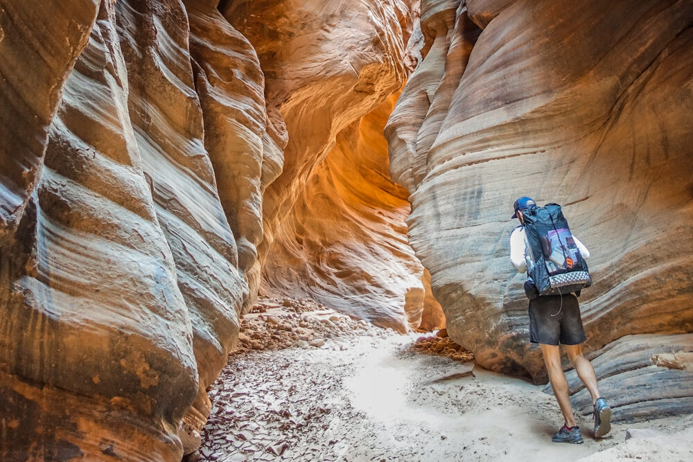 a hiker wearing a large backpack meanders through a tunnel like slot canyon section