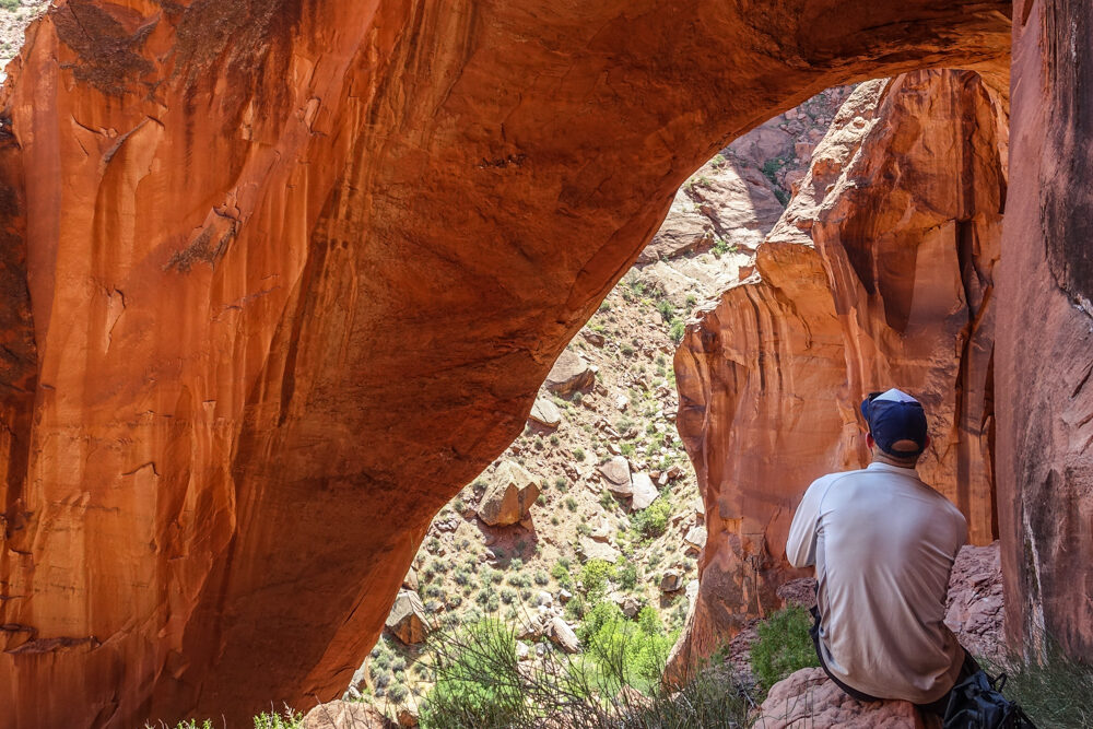 a hiker seated below a natural bridge or arch made of sandstone with a sunlit boulder field in view through it