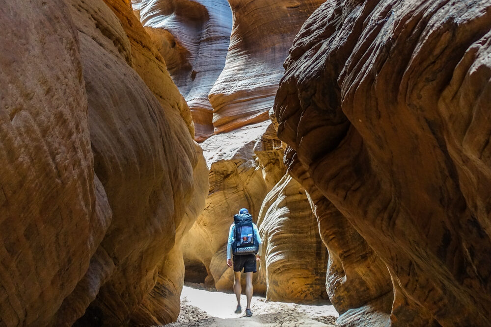 a backpacker heads toward a sunlit section of a slot canyon