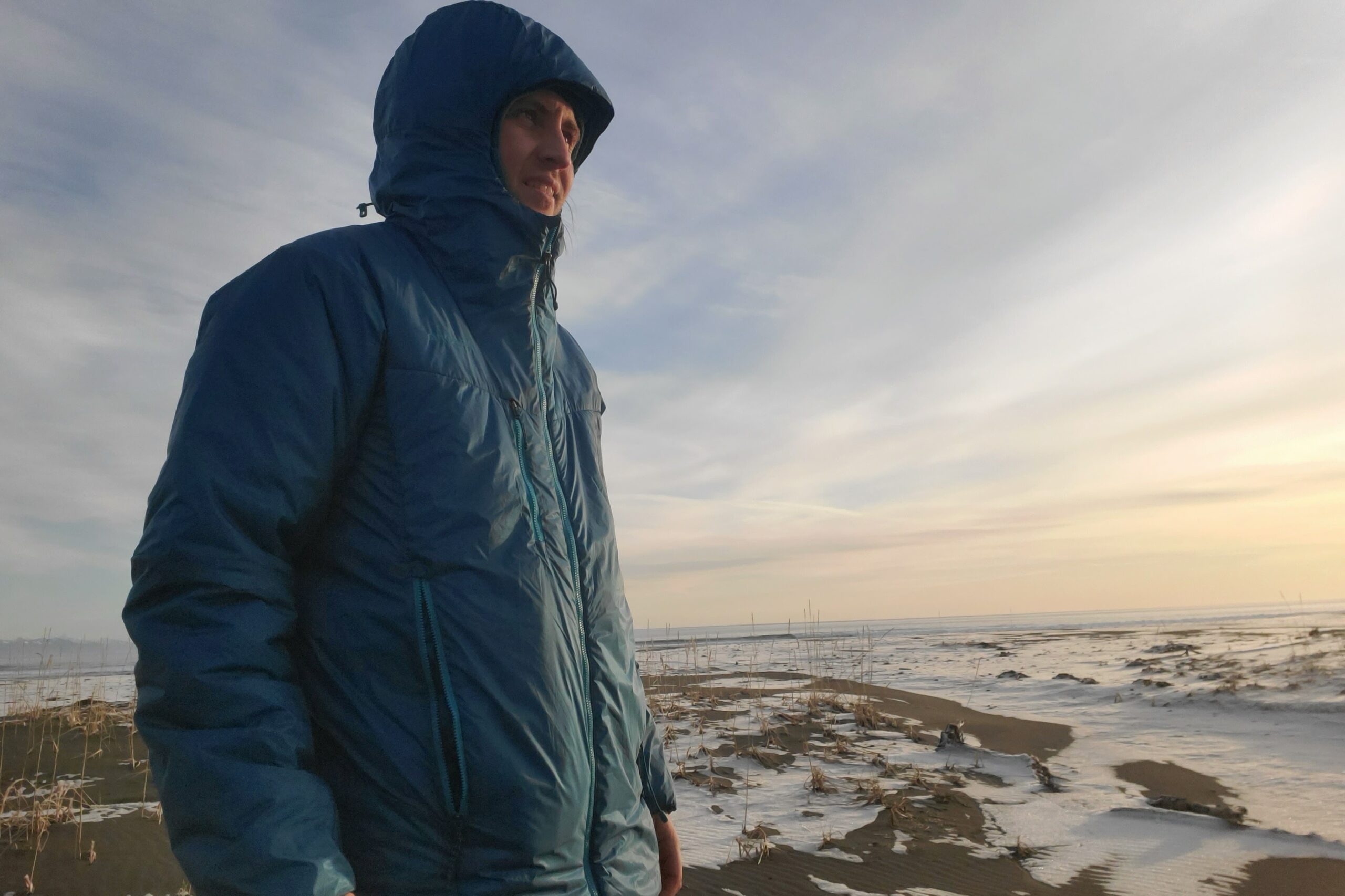 A man stands at sunset on a beach with his hood up.