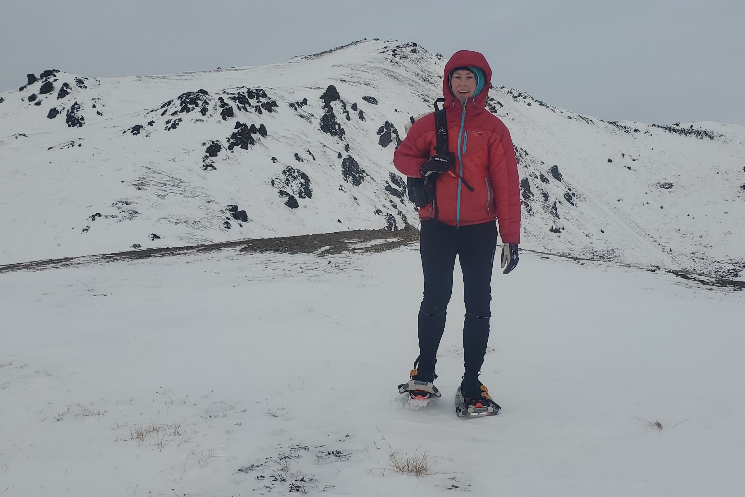 A woman walks along a snowy ridge with a mountain in the background.