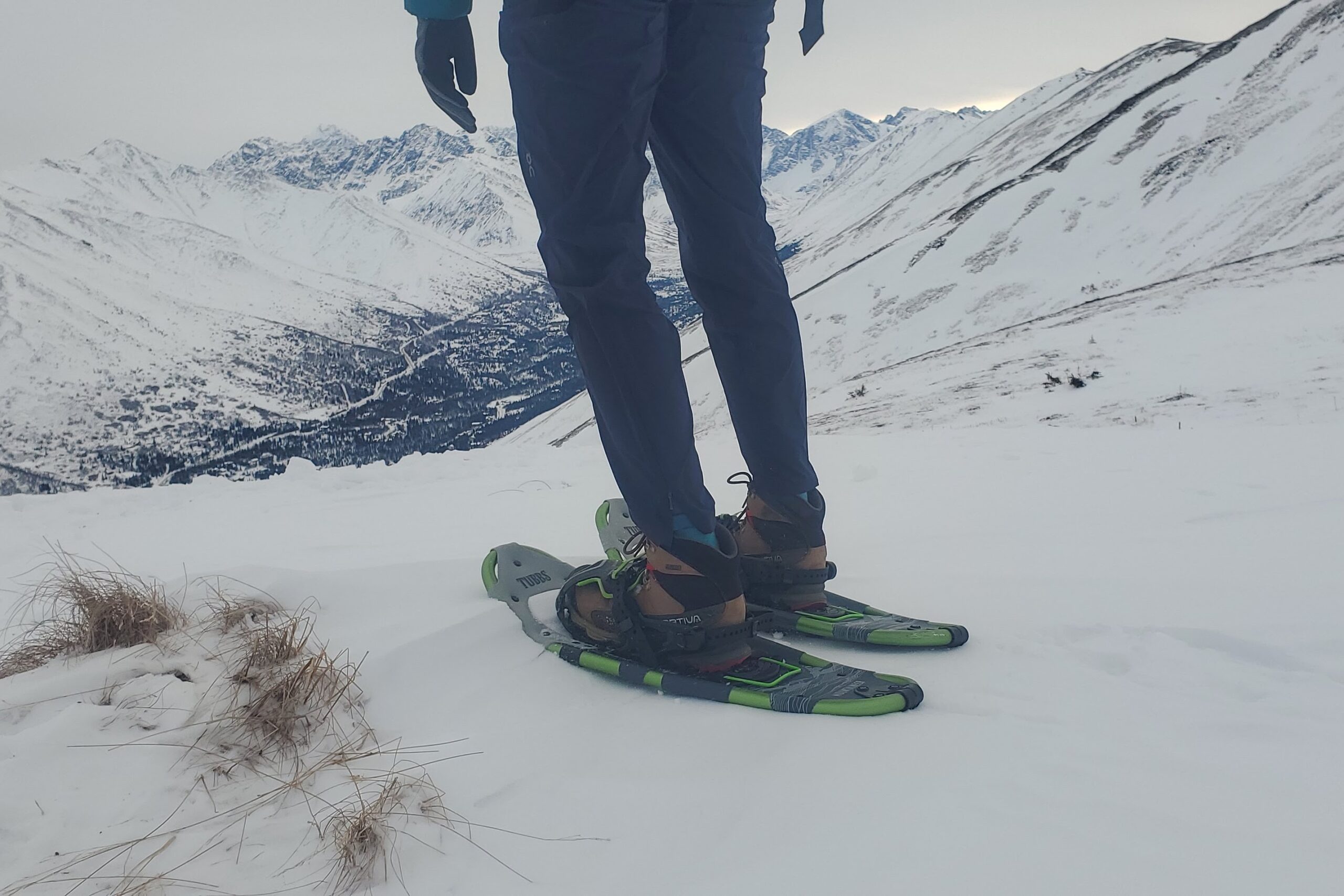 A person stands in front of a mountain view wearing snowshoes