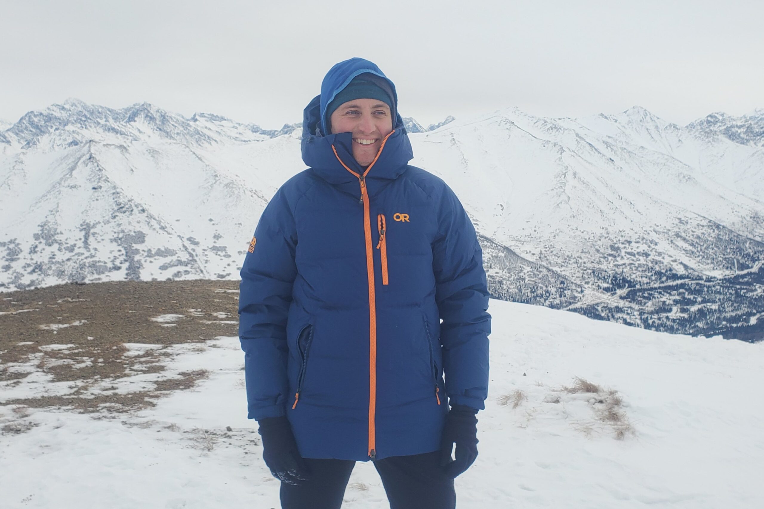 A man stands in a blue down jacket in front of a mountain landscape.