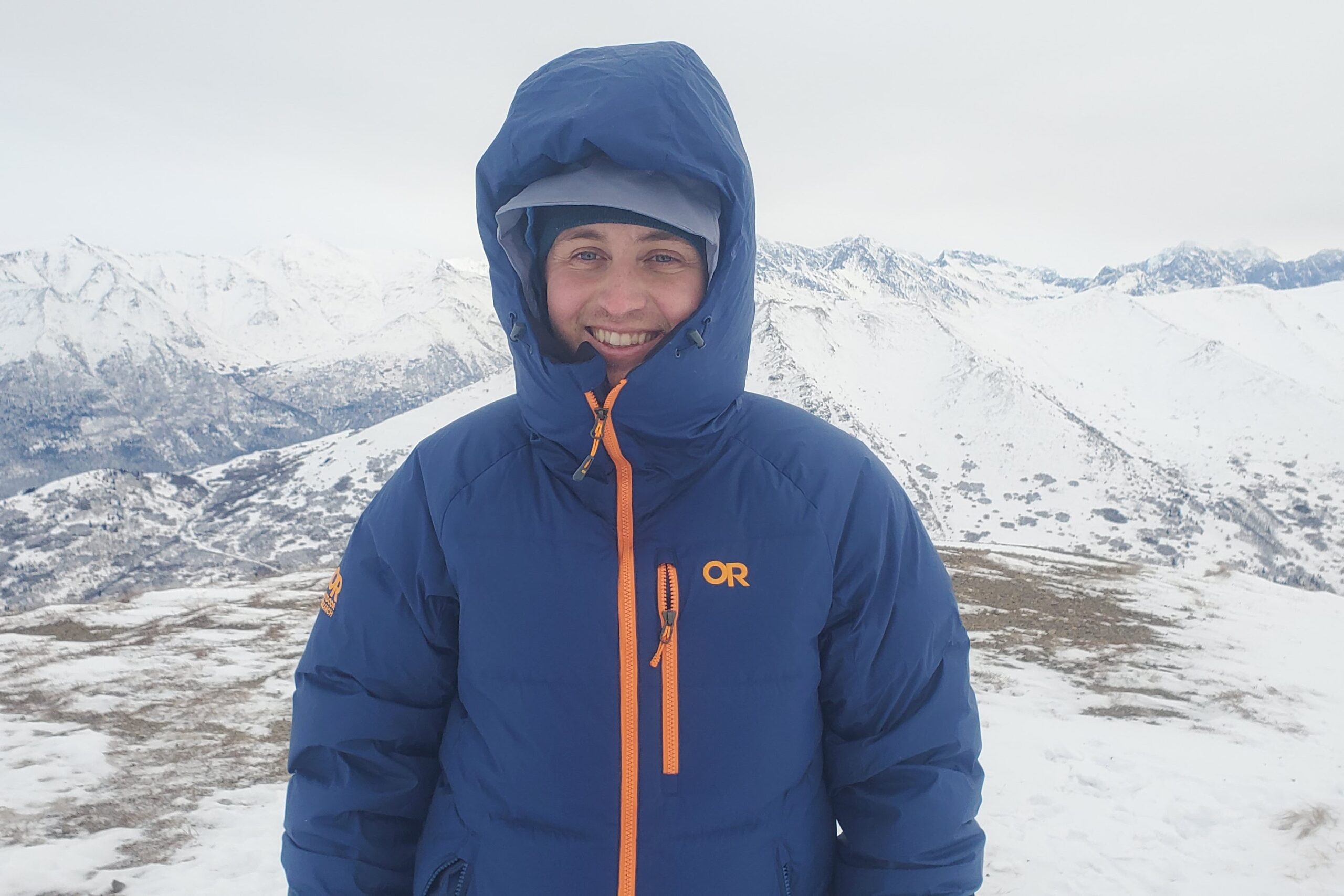 A man stands on a mountain ridge with snowy mountains in the background