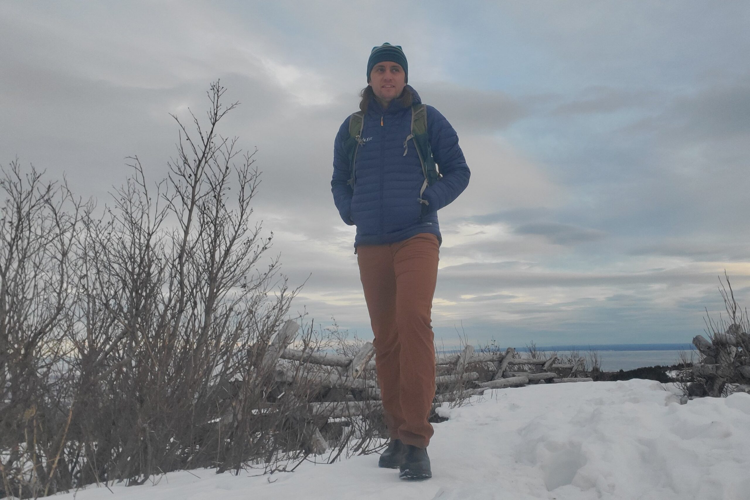 A man walks up a snowy trail with the ocean in the background.