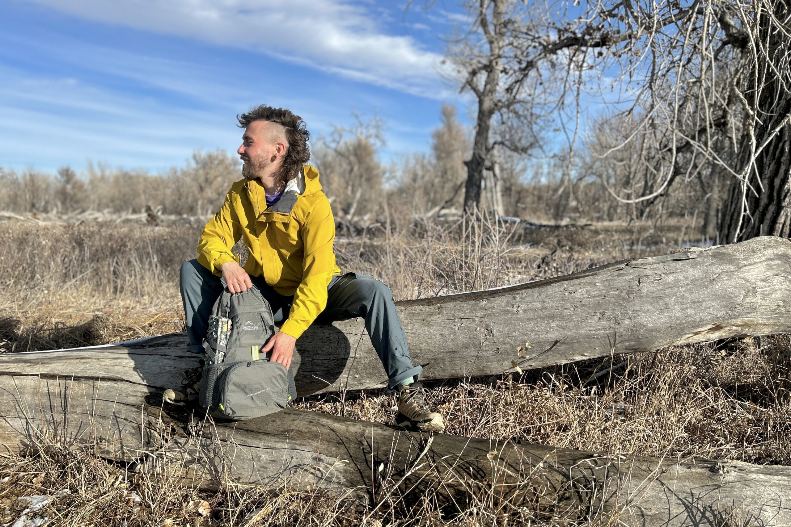 A man sits on a log and digs through a backpack in a wintry forest landscape on a sunny day.