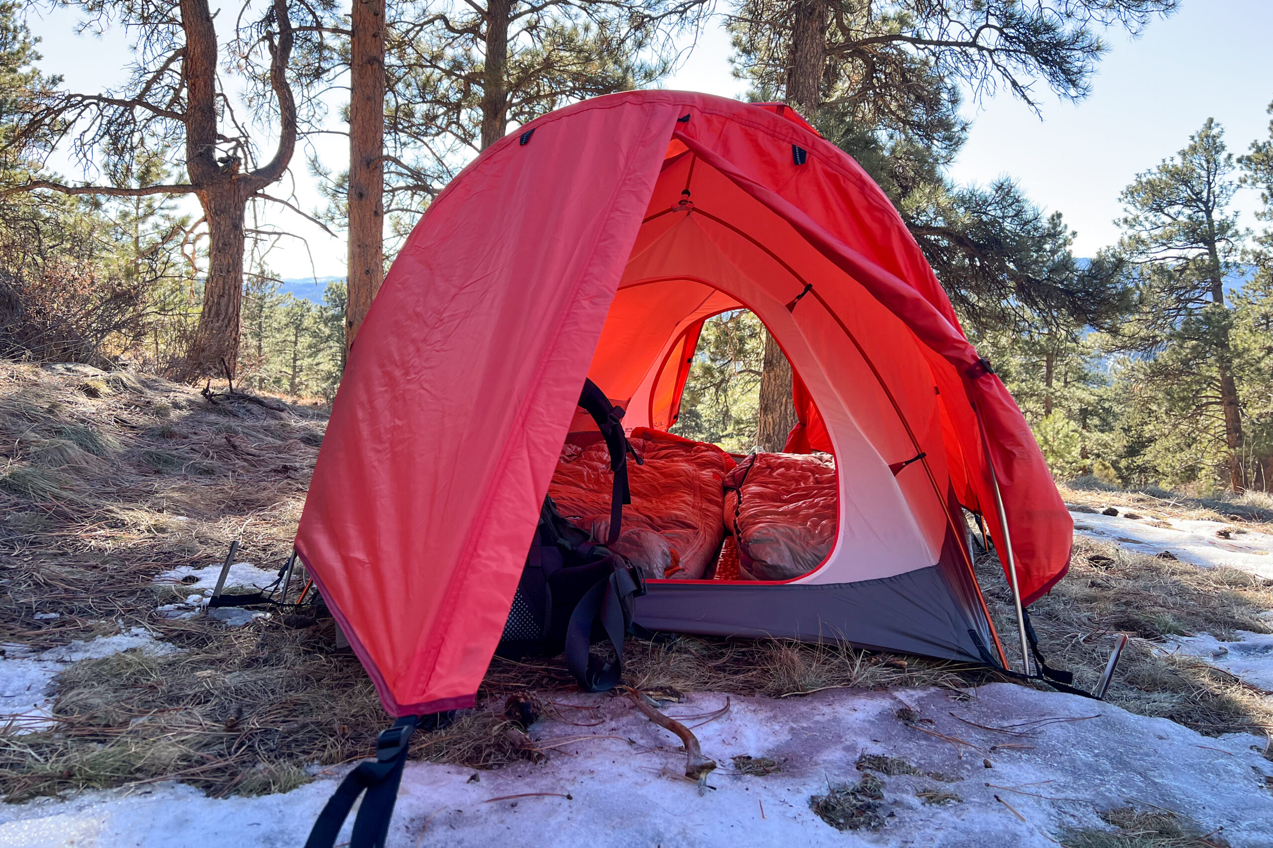 A close-up front view of the Alps Mountaineering Tasmanian 2 tent with the door open, revealing two sleeping bags inside. The red rainfly provides shelter over the entrance.