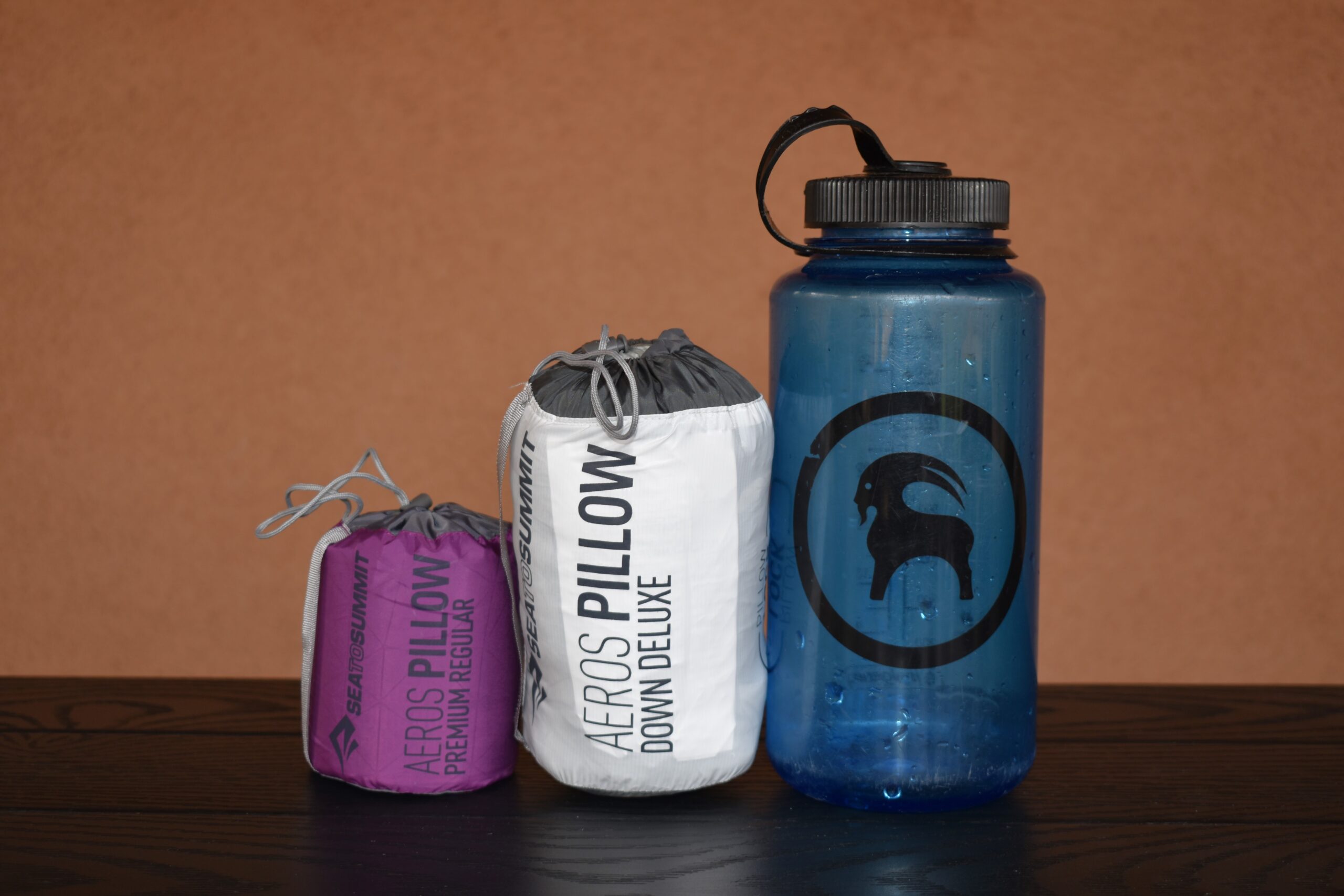 2 packed camping pillows side by side next to a 1L Nalgene water bottle. The Sea to Summit Aeros Premium Pillow is much smaller than the Aeros Down Deluxe Pillow