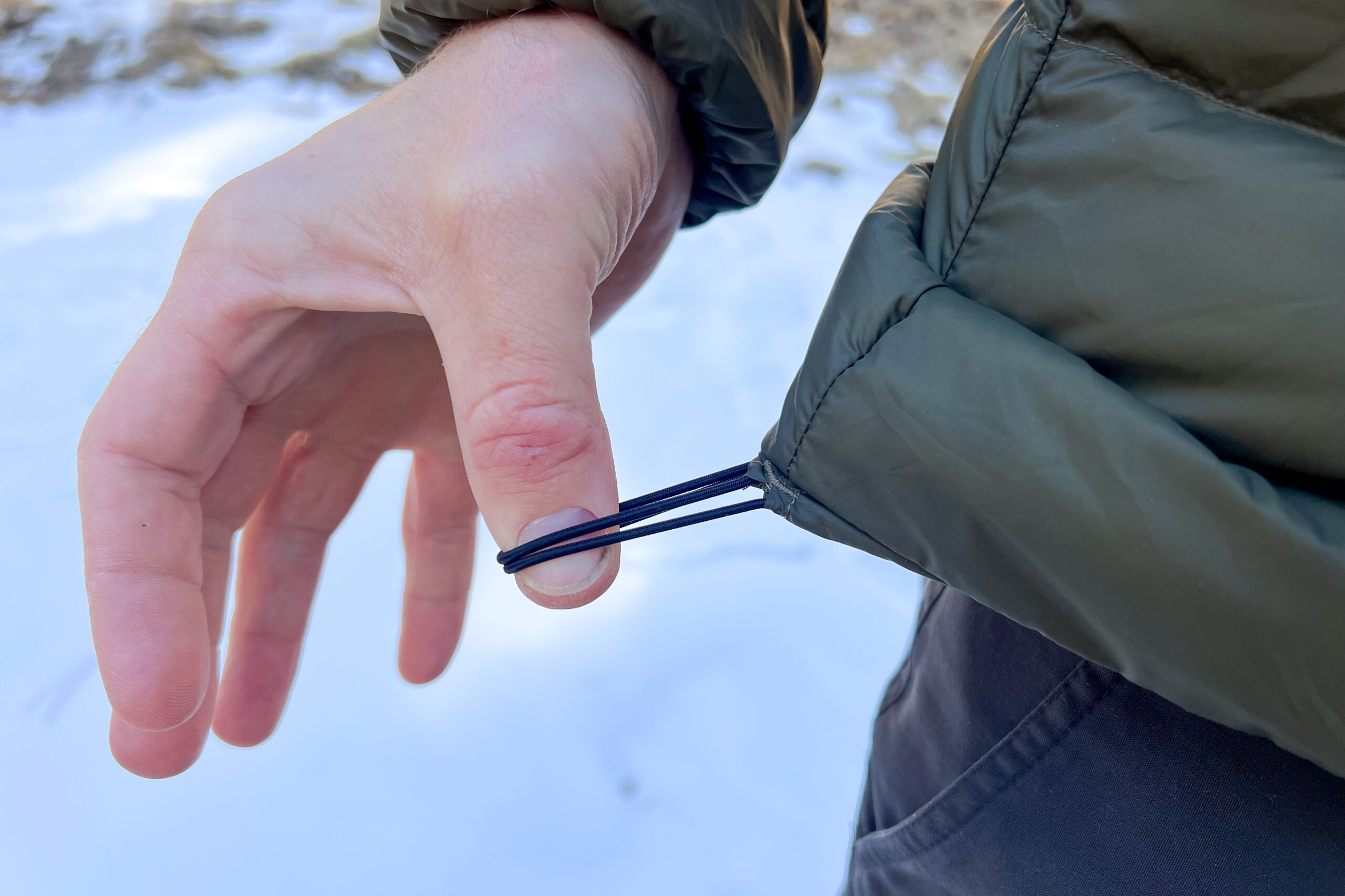 Close-up of a person pulling the adjustable drawcord on the hem of the Arc'teryx Cerium Hoody, with a snowy background visible.